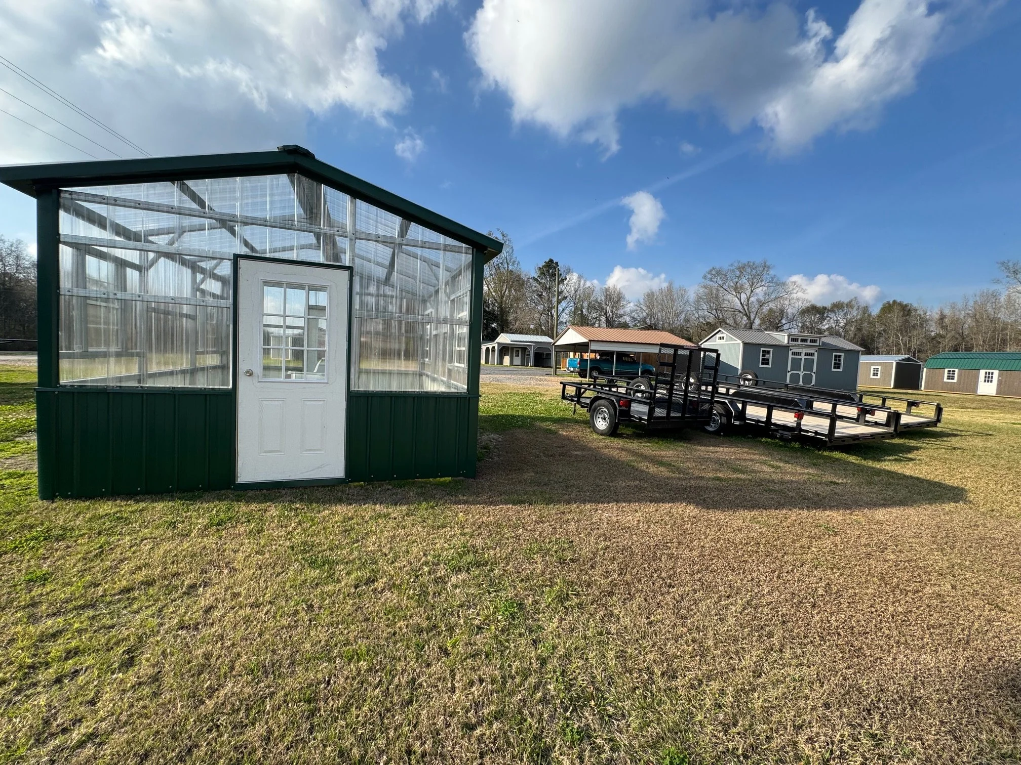 Empty greenhouse with white door, green lower panels and transparent walls in a grassy field, with a flatbed trailer nearby, against a background of small buildings and a blue sky with clouds.