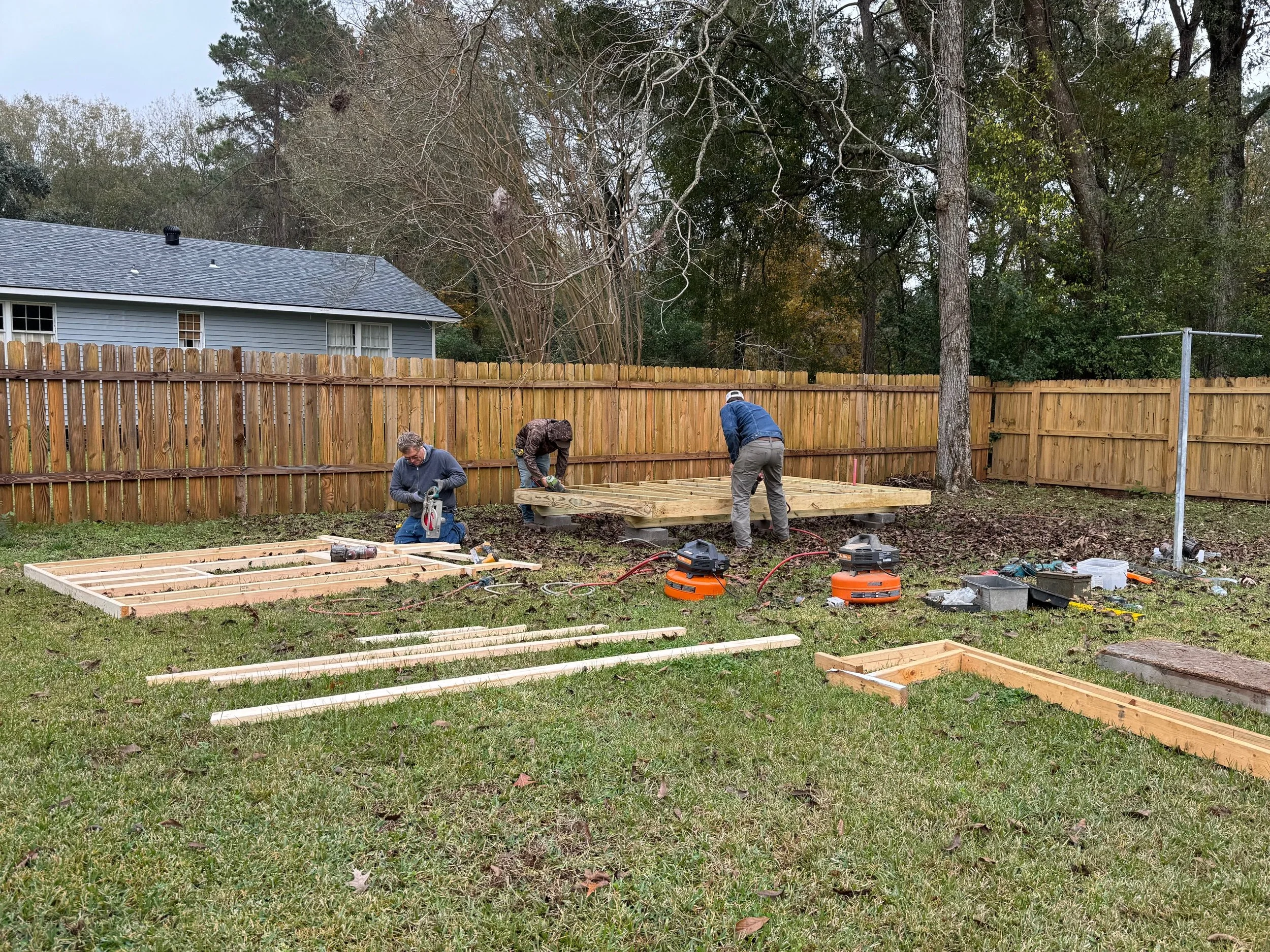 Three people building a wooden deck in a backyard, working with tools and wood framing on the grass.