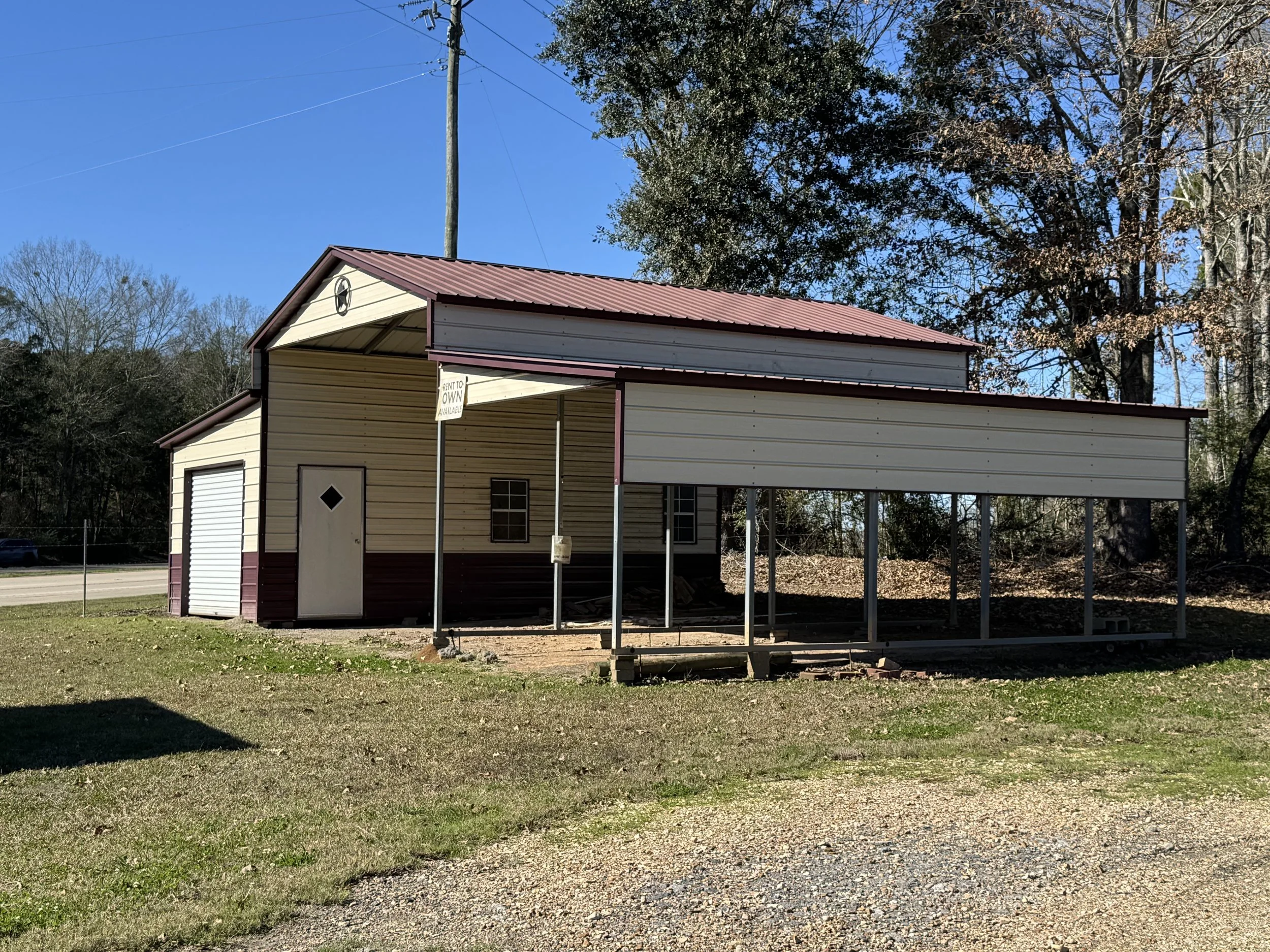 A small beige and maroon building with a garage door and a side door, with some framing for a future extension, situated on a grassy area under a clear blue sky.