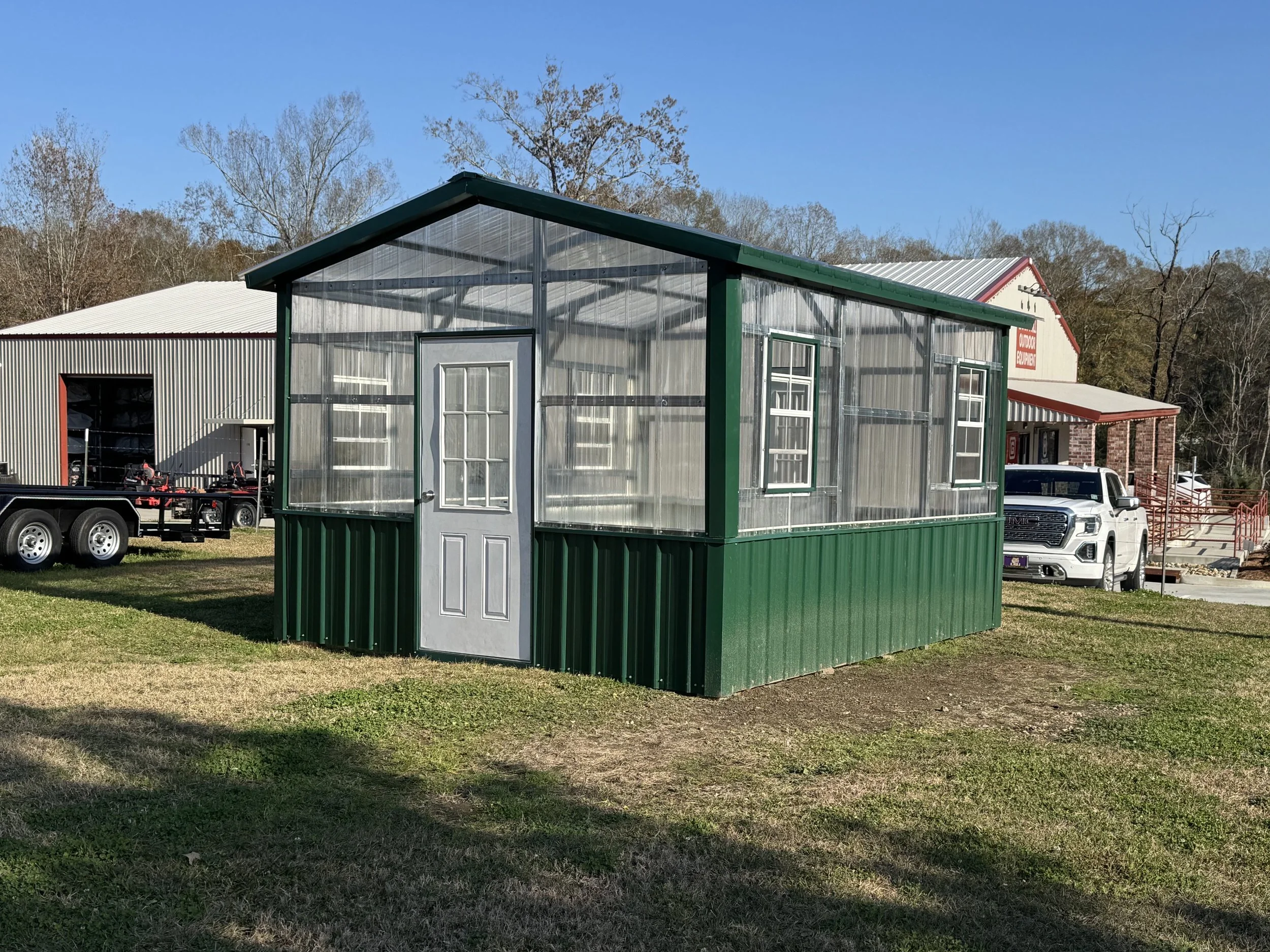 A small greenhouse with a metal door and windows, painted green, sit on a grassy area with trees in the background and a clear blue sky overhead.