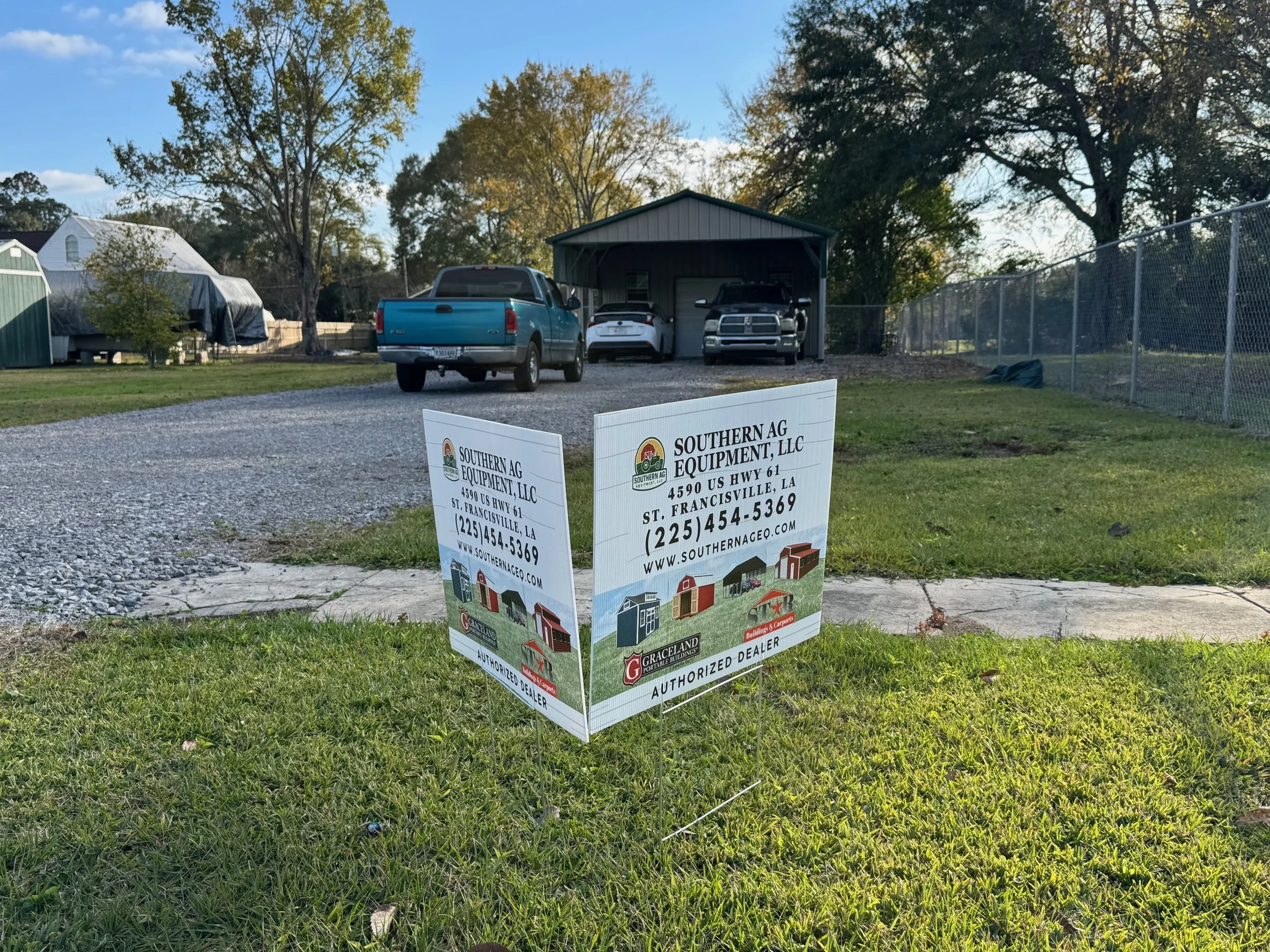 A yard with a gravel driveway leading to a garage, with vehicles parked outside, including a blue pickup truck, and a white car. There is a chain-link fence on the right, trees in the background, and two signs on the grass promoting Southern Ag Equip