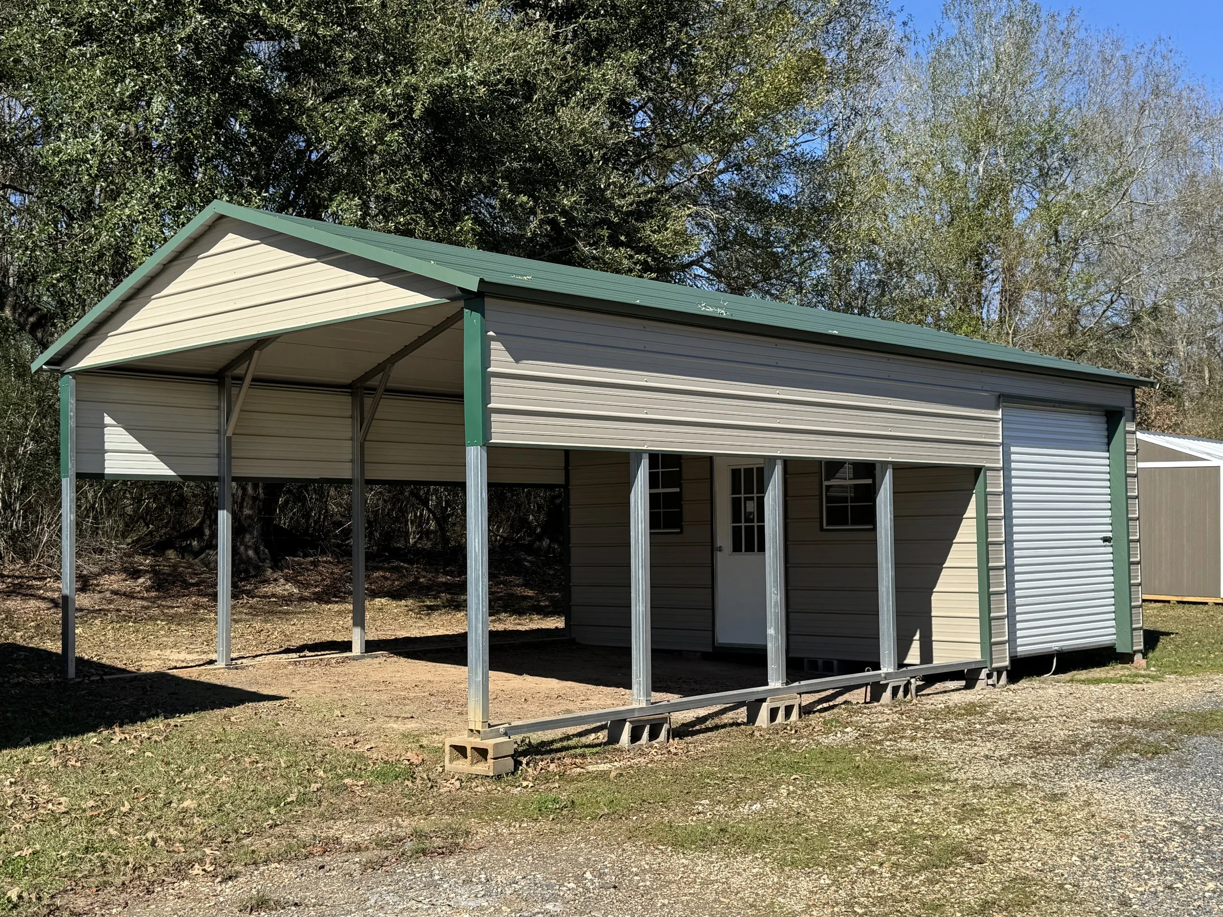 A metal carport structure with a green roof and beige siding, featuring a closed roll-up door and two small windows, situated on a grassy area with trees in the background.