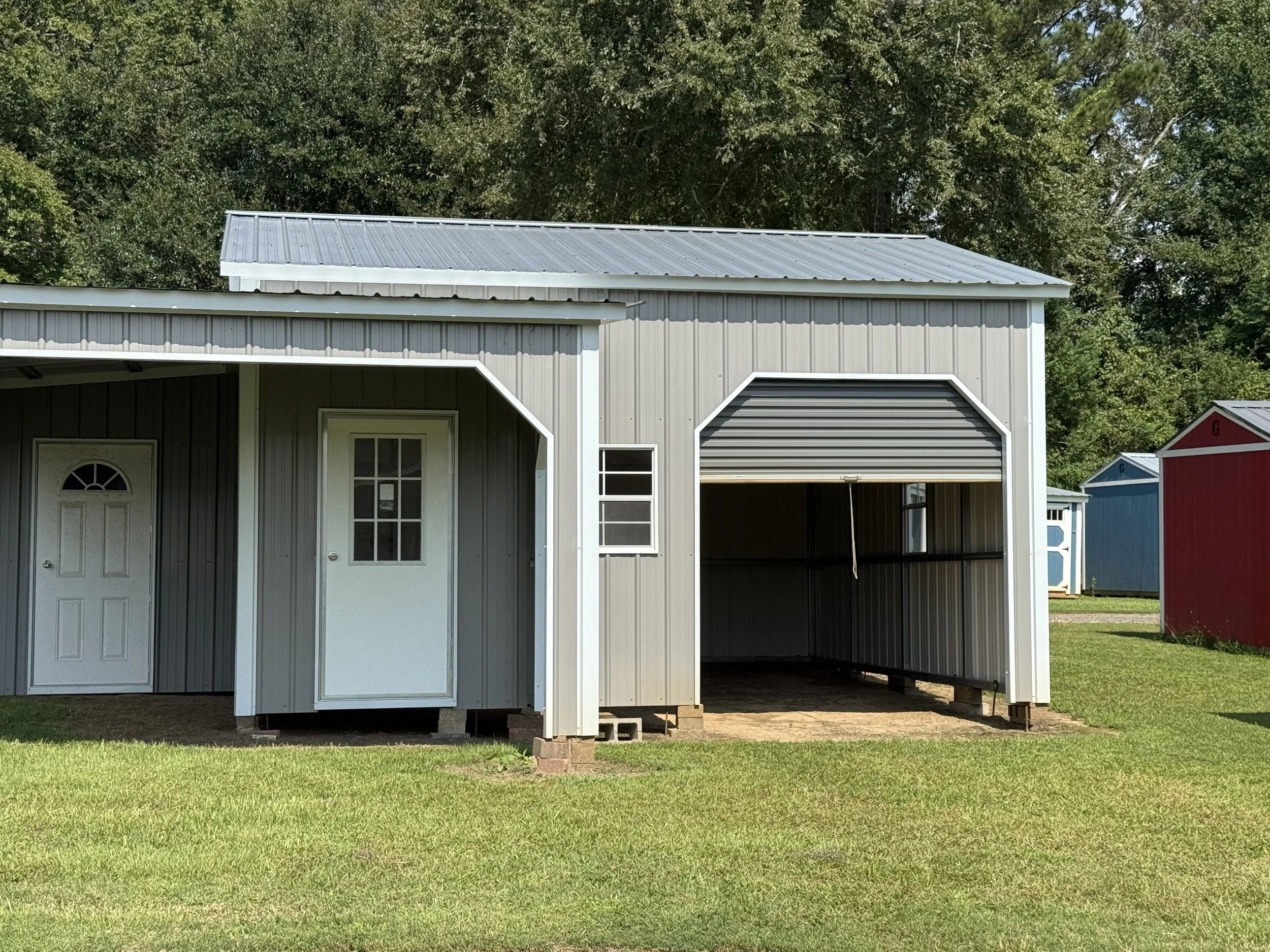 Gray metal shed with a partially open garage door and a side door, situated on grass with similar smaller sheds in the background, under a tree line.