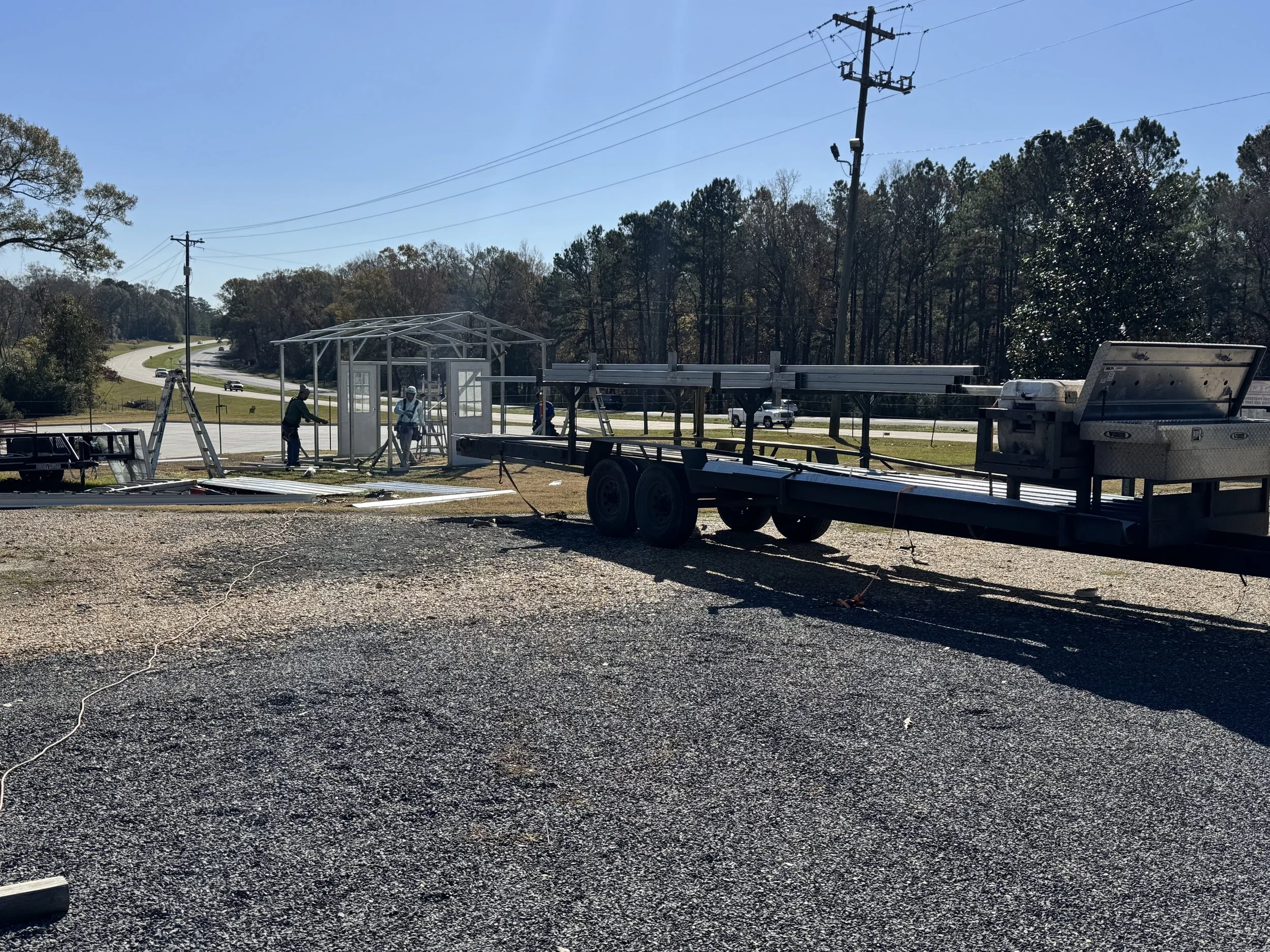 Construction workers assembling a small structure on a trailer with power lines and trees in the background on a clear, sunny day.