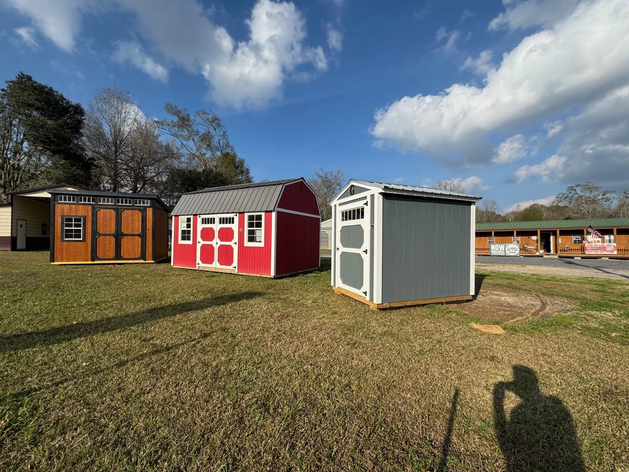 Three small storage sheds in different colors and styles are placed on a grassy area under a partly cloudy sky. A building with a porch is visible in the background on the right.