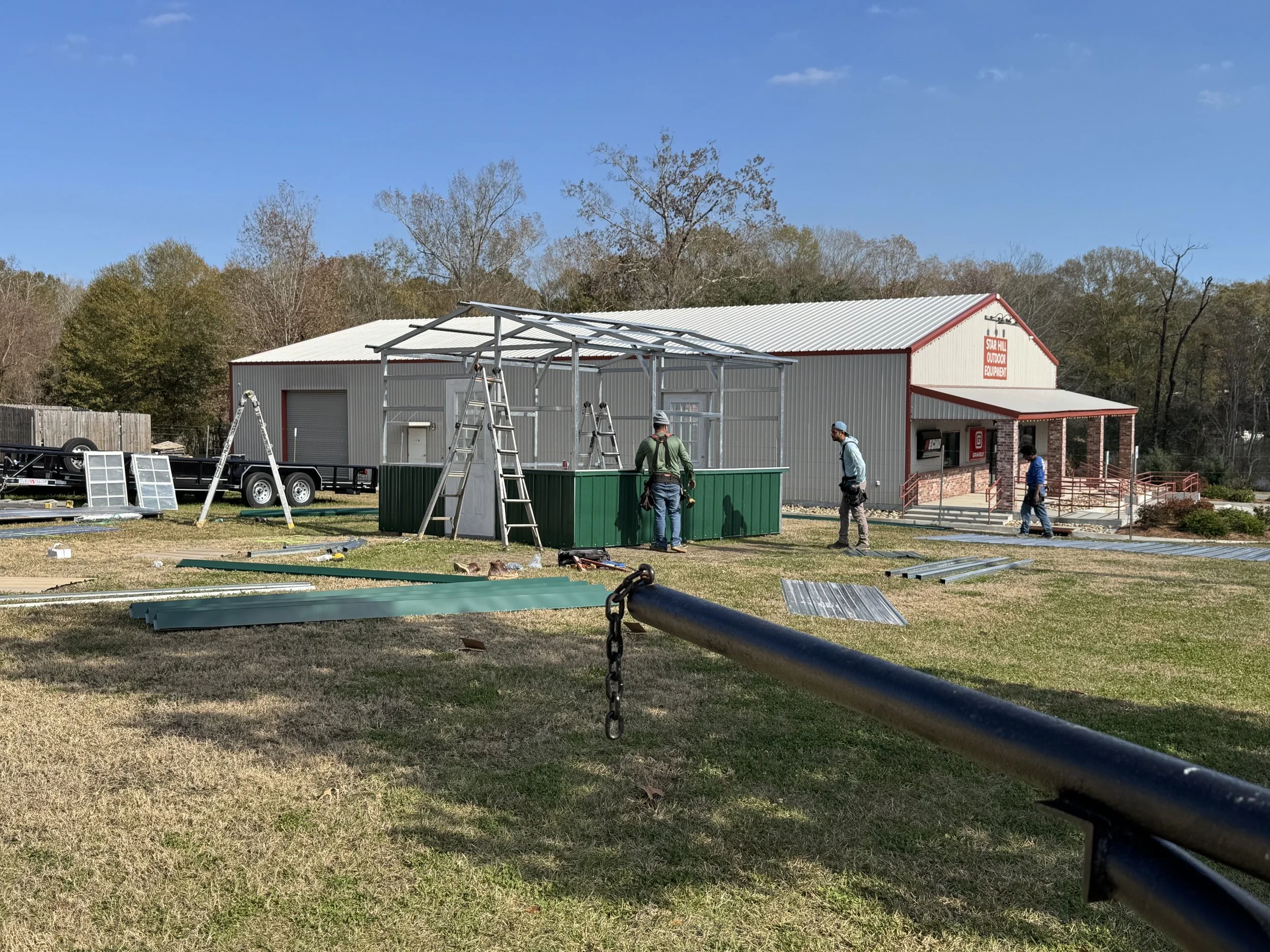 People working on assembling a small outdoor building with metal framing in front of a larger barn-style building with a sign that reads 'Star Hill Outdoor Equipment,' surrounded by trees and grass on a sunny day.