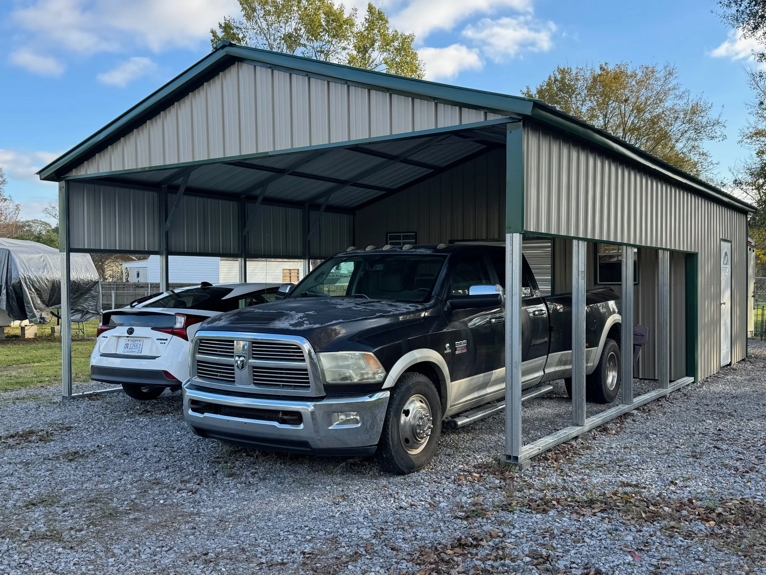 A black Dodge Ram pickup truck parked in a metal carport with a white Tesla behind it. The carport is on gravel and surrounded by trees and a fenced yard.