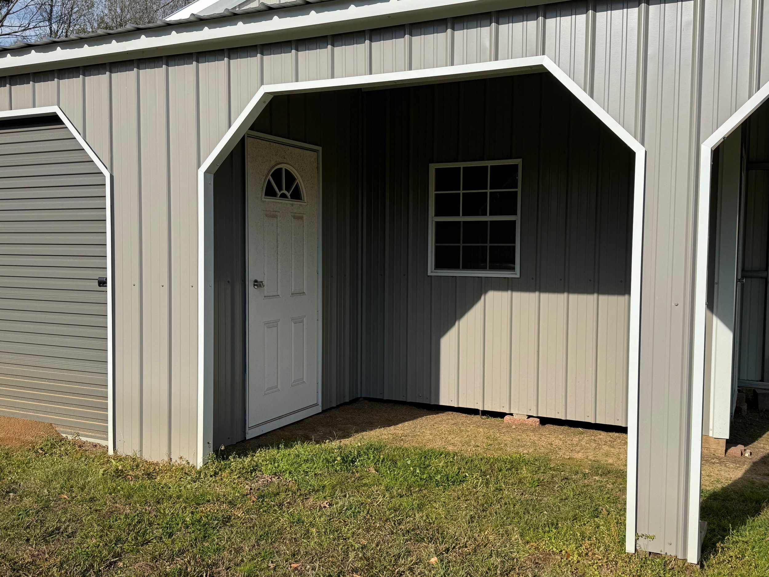 Small metal shed with a white door and a window, situated on grass with a patch of dirt underneath.