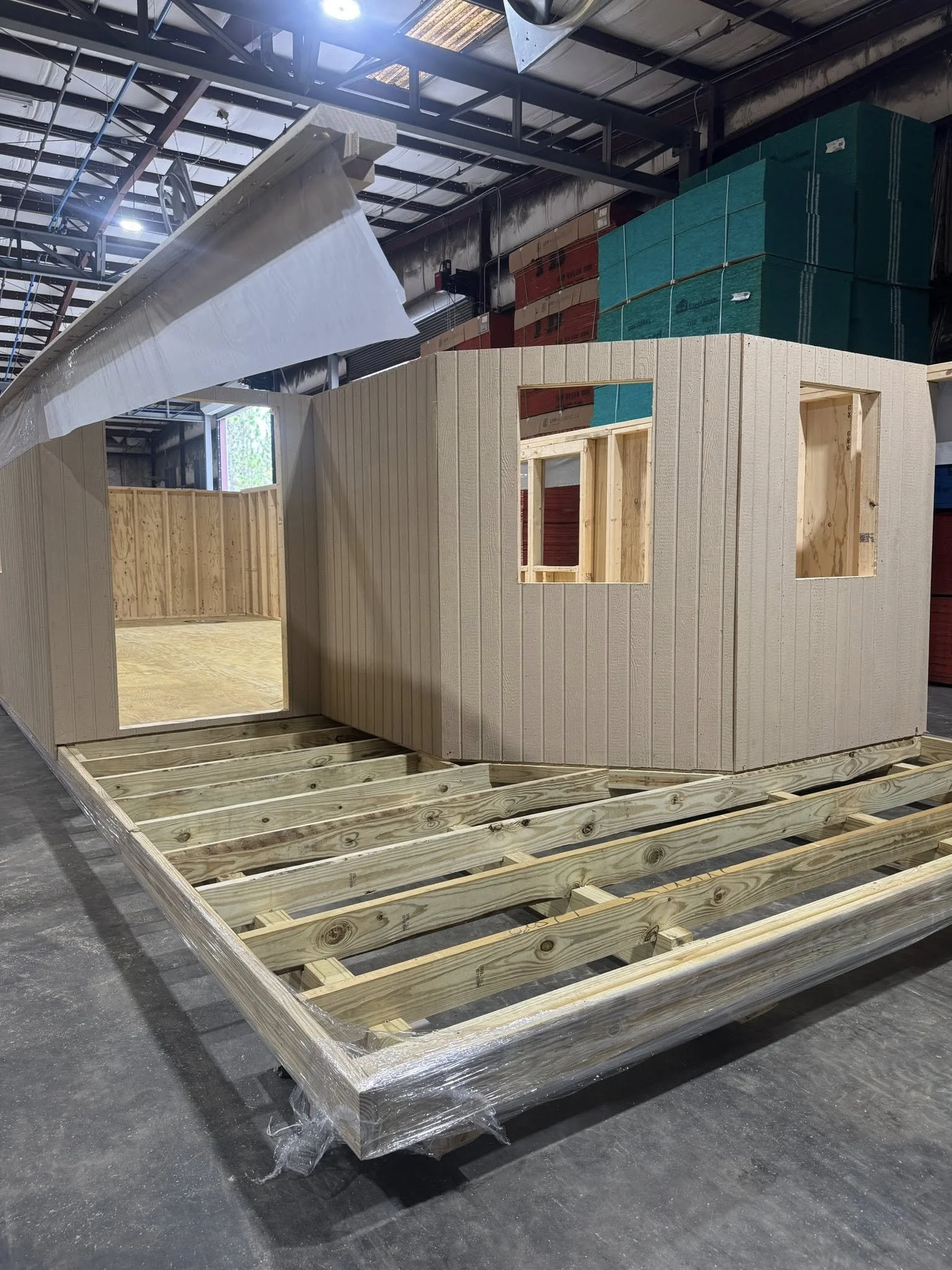 A partially constructed wooden building on a framing platform inside a warehouse. The structure has walls with vertical siding and multiple window openings, with the roof partially installed.