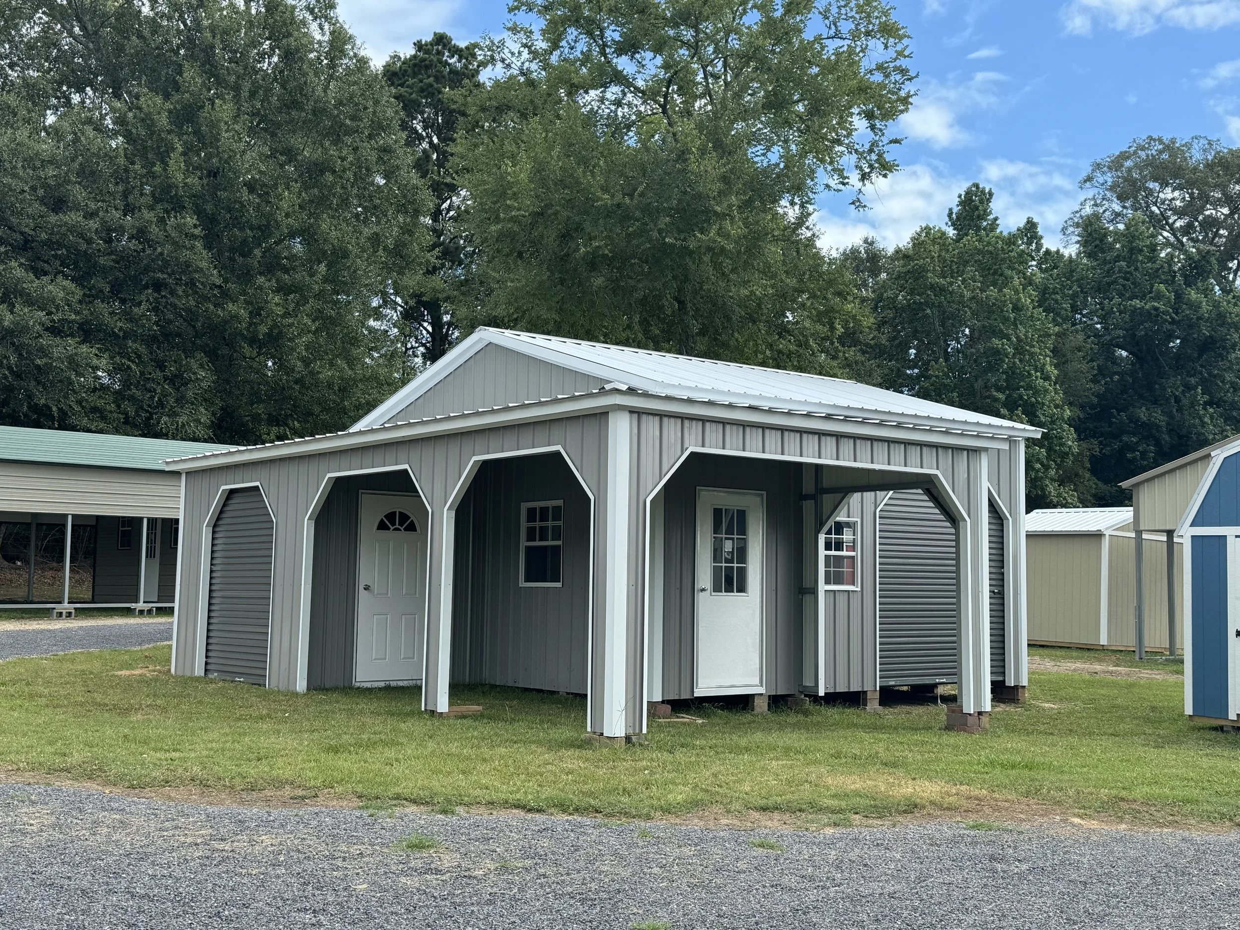 Gray storage shed with white trim and a metal roof, featuring two doors and two windows, situated on a grassy area with a gravel path nearby, surrounded by trees.