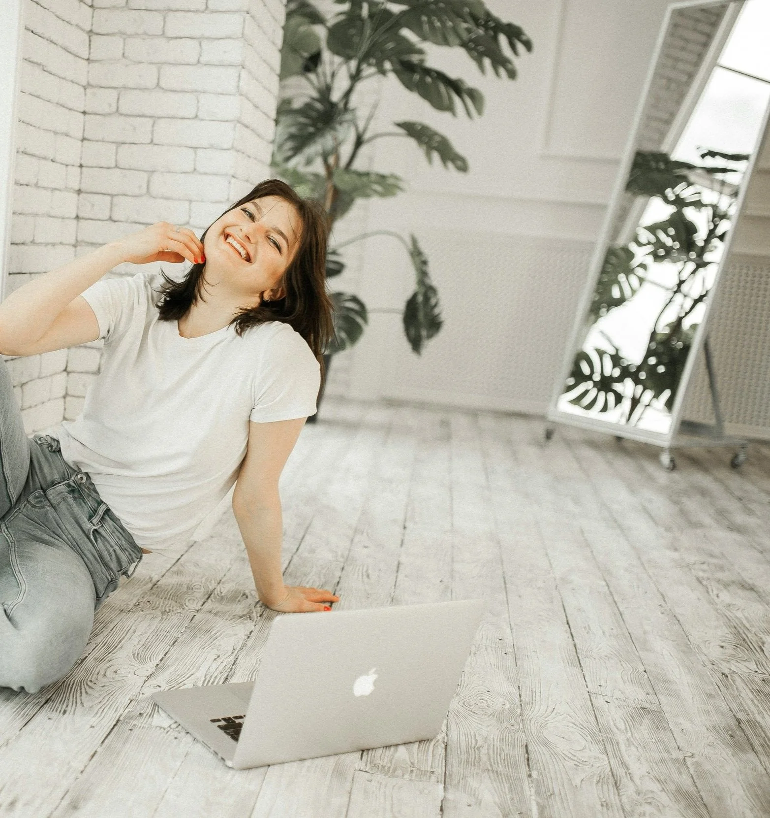 A woman sitting on the floor with a MacBook laptop, smiling and touching her face, in a bright room with large mirror, plants, and white brick wall.