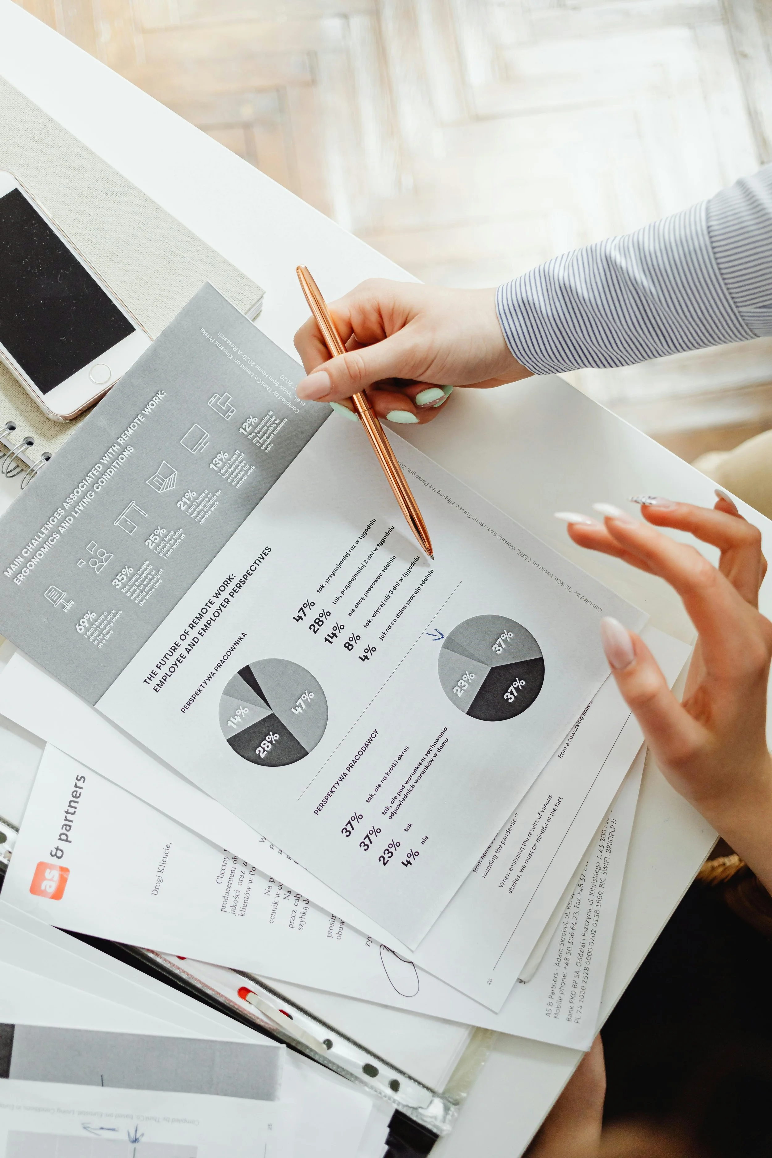 A person holding a pen and pointing at a document with pie charts and data, sitting at a white table with a smartphone and papers.