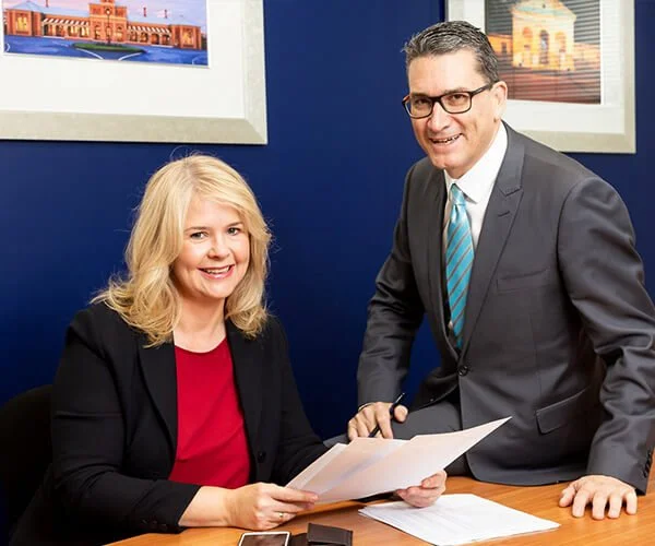 A woman sitting at a desk holding papers and a man standing next to her in a gray suit, both smiling, in an office with framed pictures of bridges on the wall behind them.