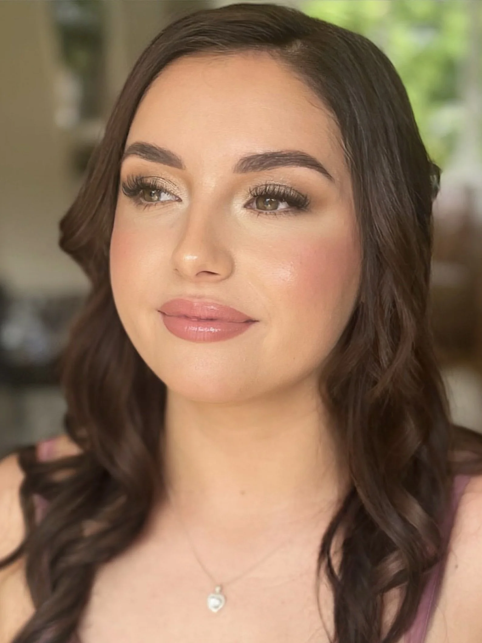 A woman with wavy brown hair and natural makeup, wearing a pink top and heart-shaped pendant necklace, looking to the side.