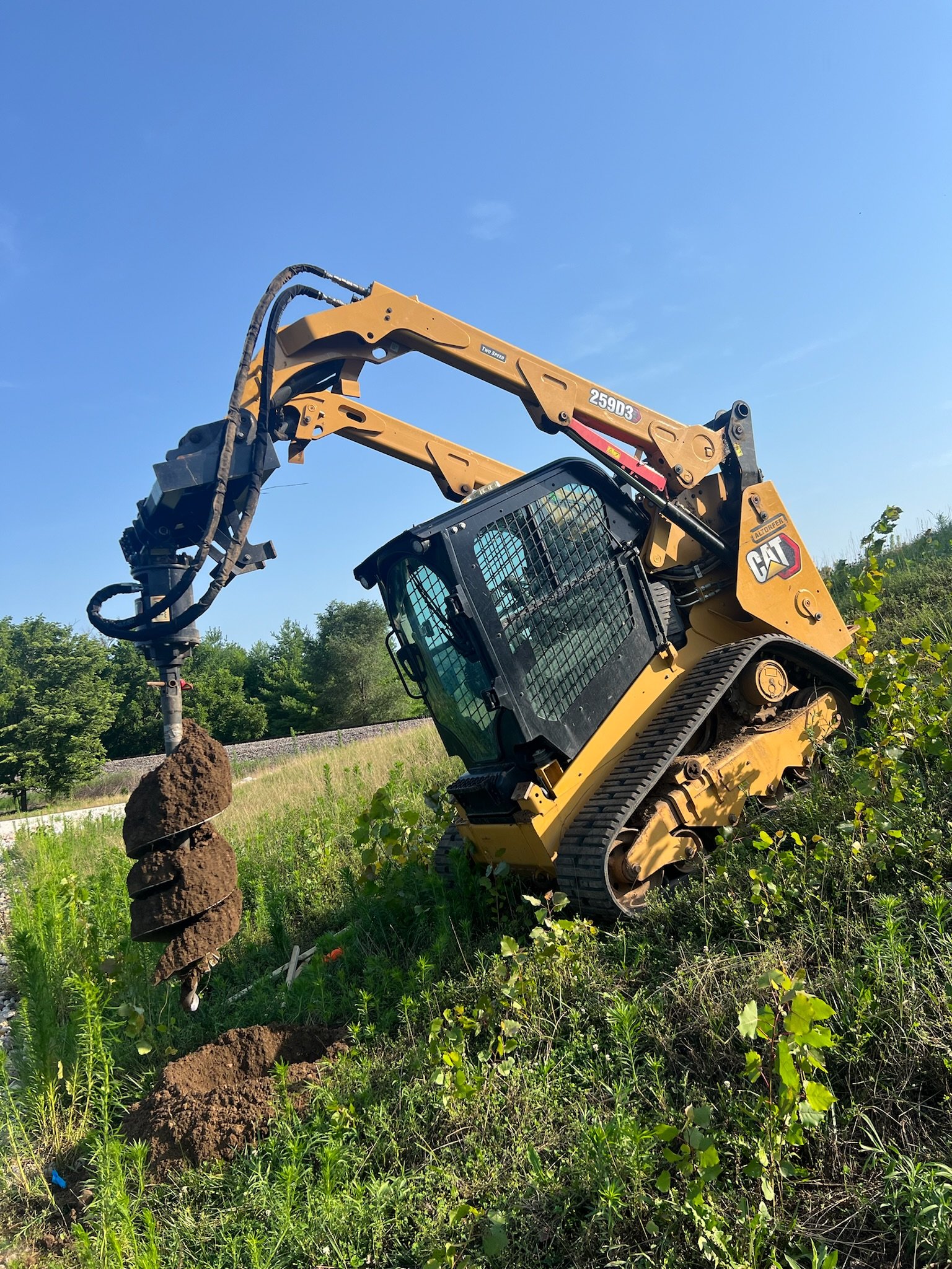 A yellow Caterpillar compact excavator digging into the ground in a grassy field under a clear blue sky.