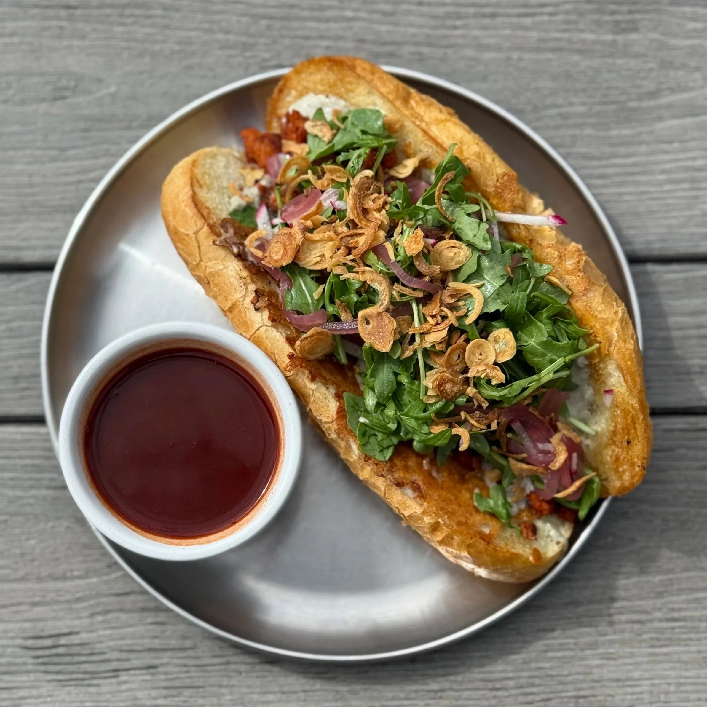 Vietnamese banh mi sandwich with fresh herbs, fried shallots, and a side of dipping sauce on a metal tray.