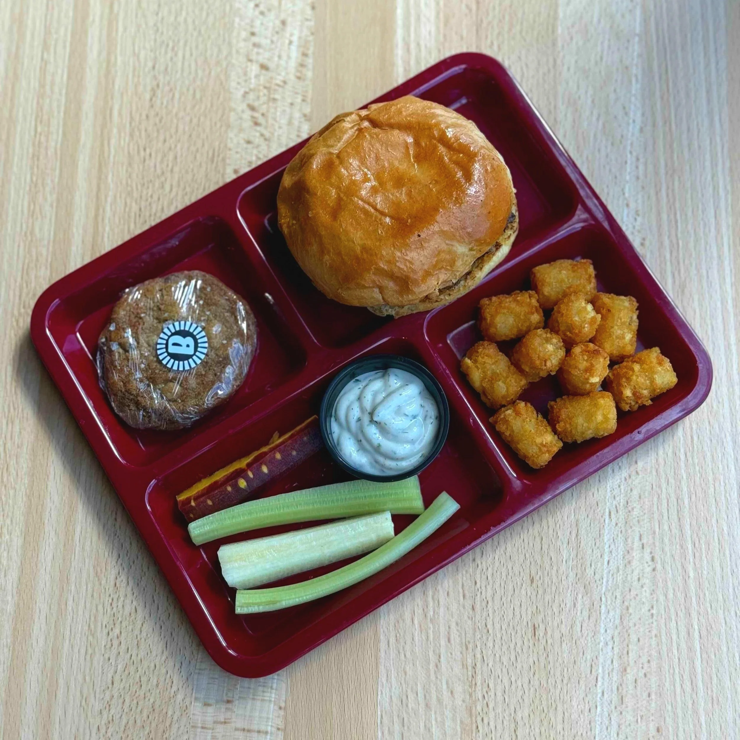 School cafeteria tray with a cheeseburger bun, chicken nuggets, a cookie, celery sticks, and ranch dip.
