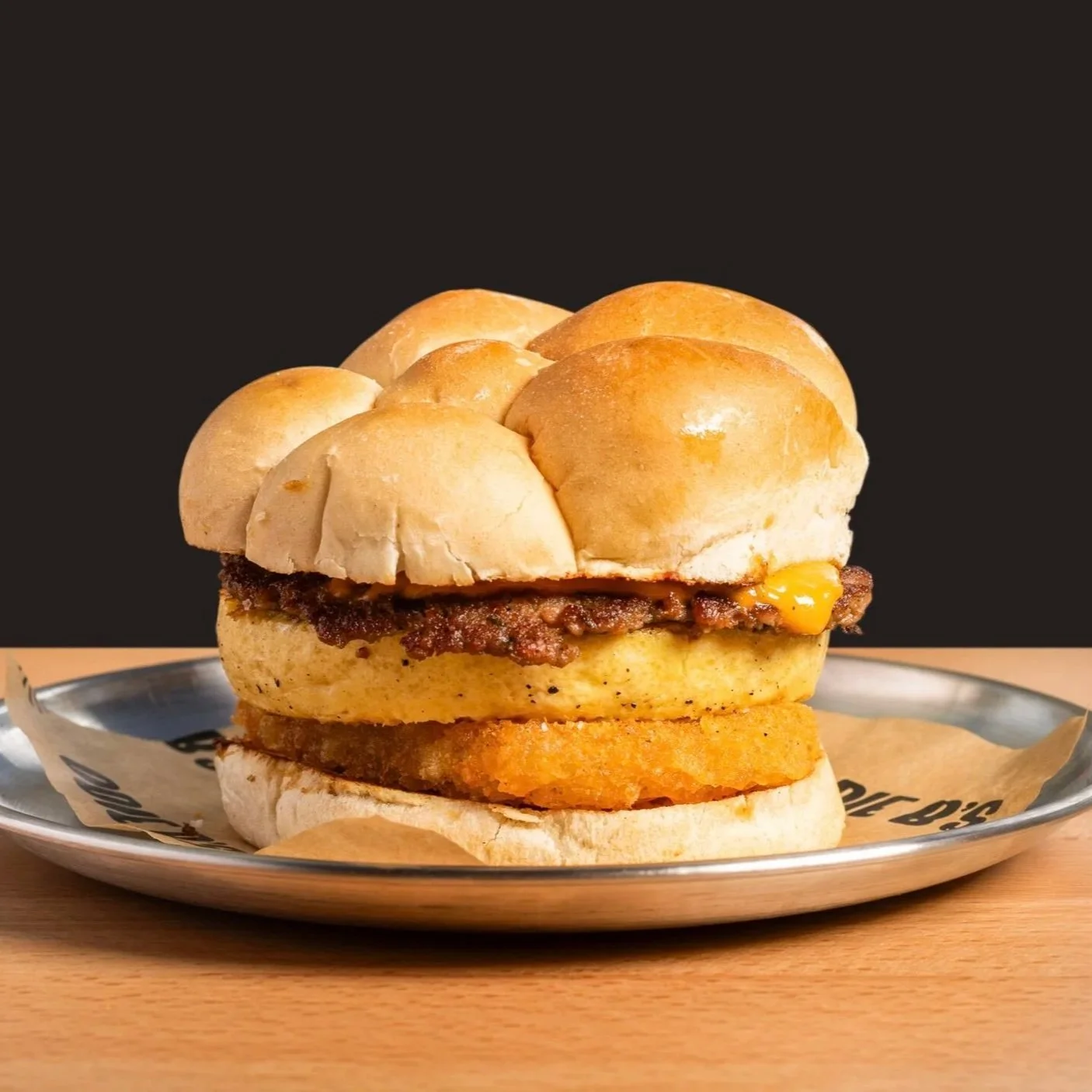 A burger with a bun top topped with a cluster of small bread rolls, a beef patty, cheddar cheese, fried onion rings, and a toasted bun bottom served on a metal tray with parchment paper, against a black background.
