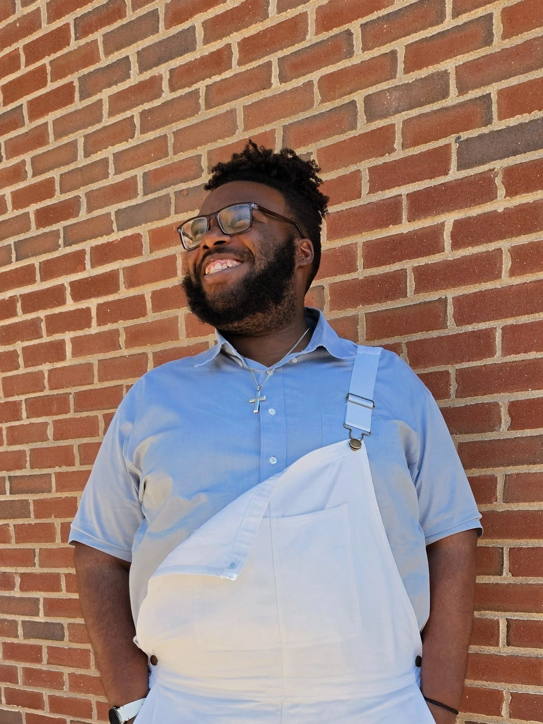 A man with glasses, a beard, and short curly hair standing against a red brick wall, smiling and wearing a light blue shirt with an apron and a necklace with a cross pendant.