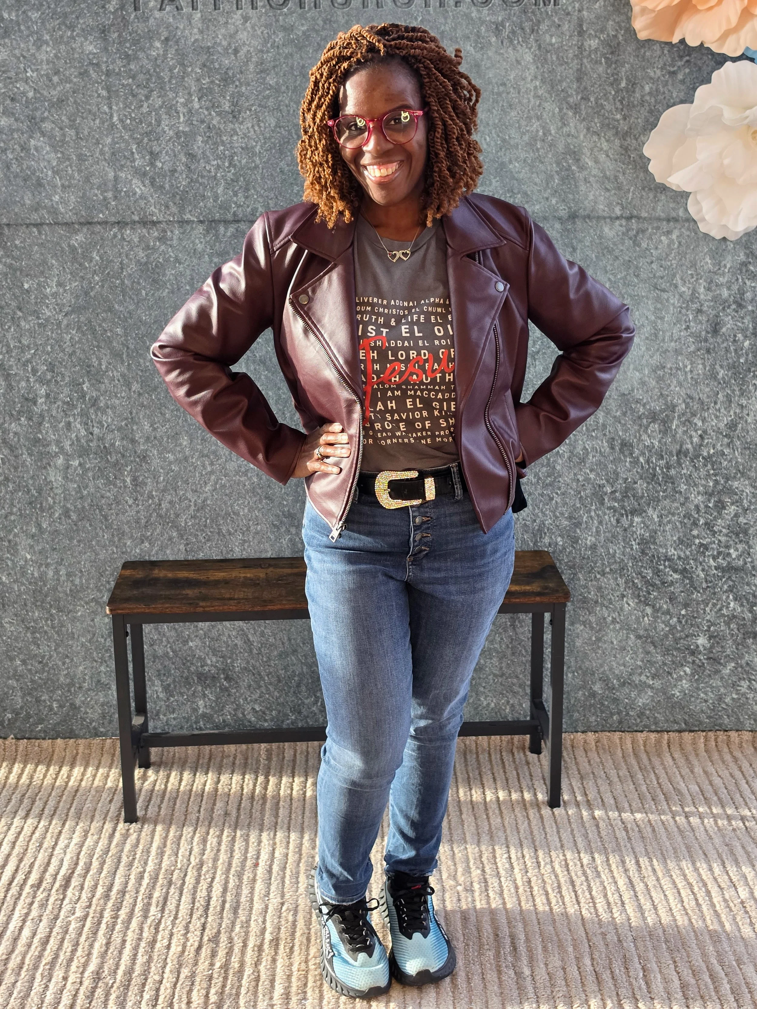 A woman with curly hair wearing glasses, a leather jacket, a graphic T-shirt, jeans, and sneakers, standing in front of a gray wall with white flowers and a bench underneath.