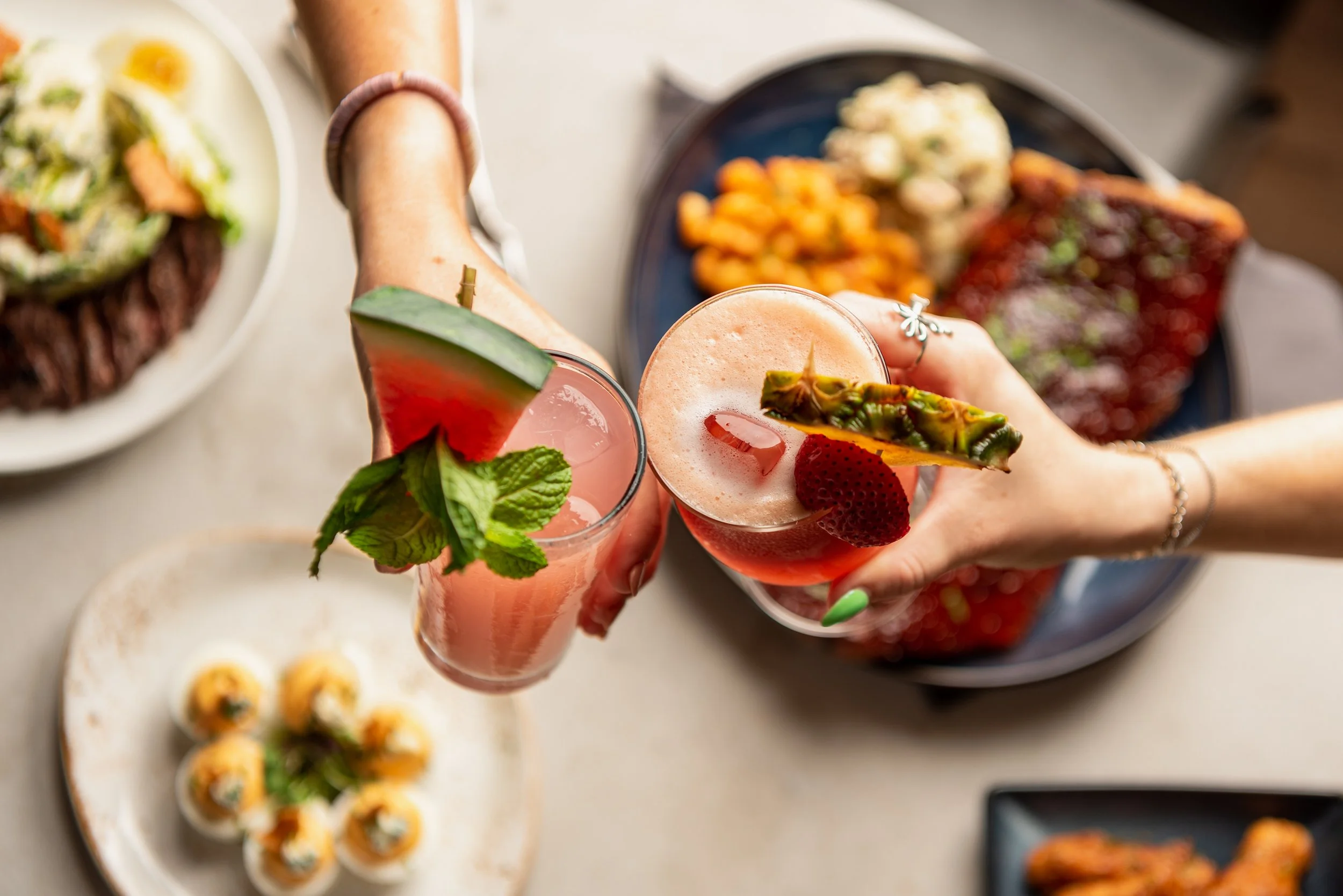 two hands toasting and clinking glasses of summery cocktails, with a background of ribs, caesar salad, and deviled eggs