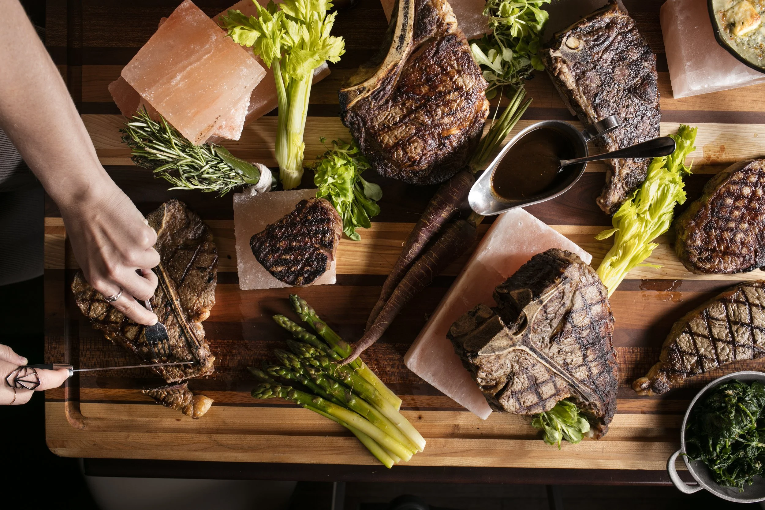 a women's hands cutting a steak on a cutting board full of dry-aged steaks, with bricks of pink himalayan salt vegetable accoutrements.