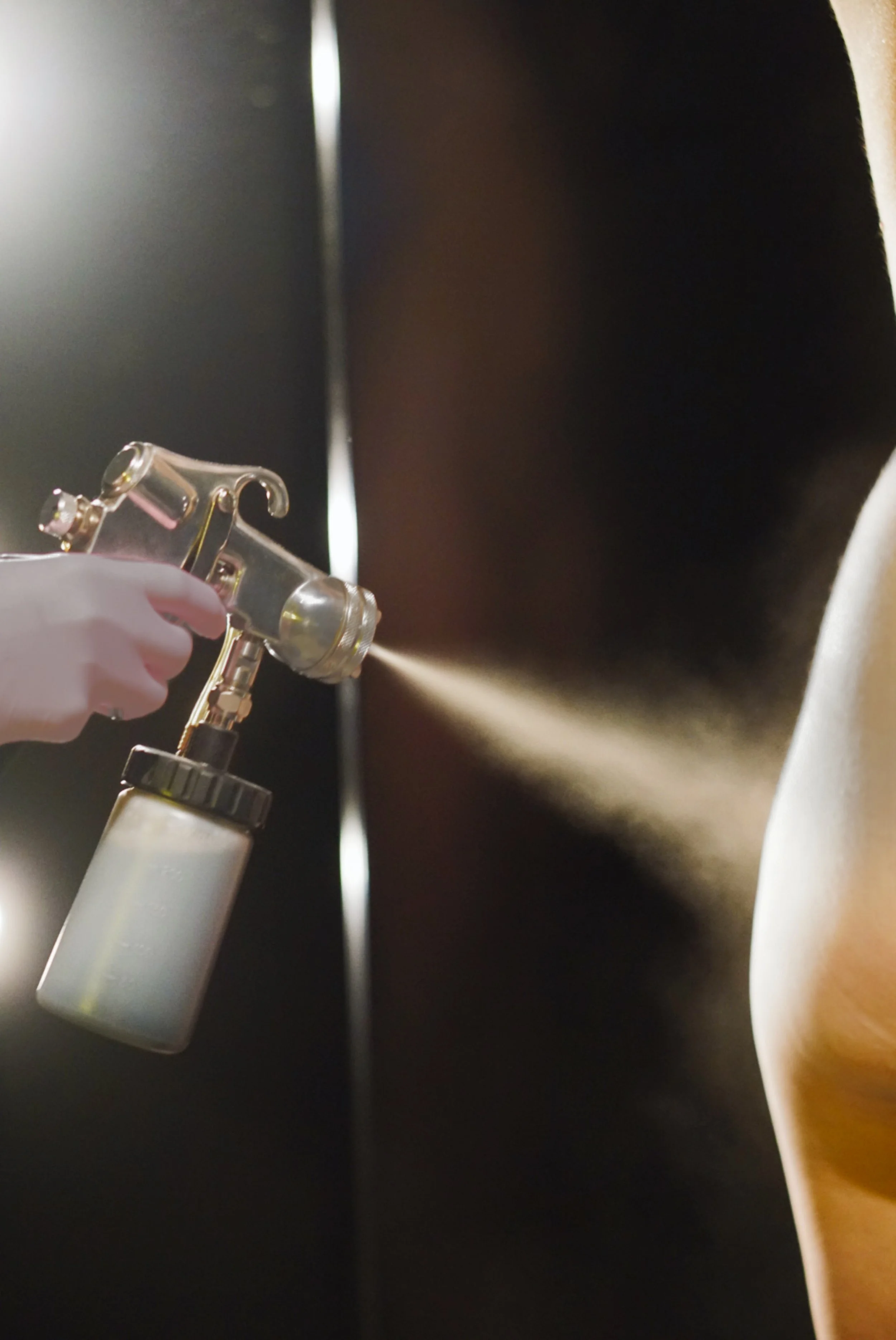 A person is using a spray gun to spray a substance onto an object. The person's hand is wearing a white glove. The background is dark with a streak of light.