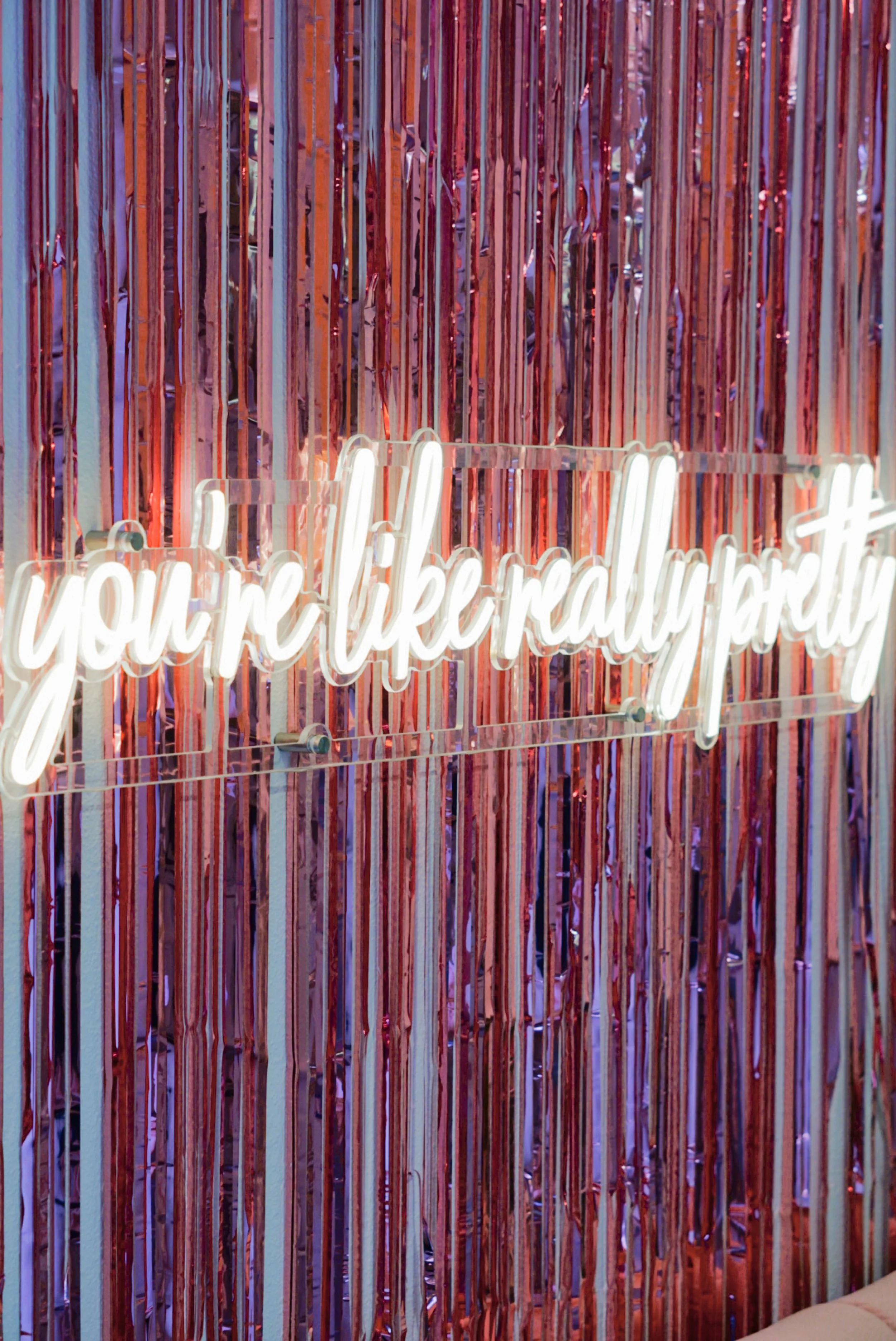 Neon sign reading 'you're like really pretty' mounted on a backdrop of vertical pink, purple, and red metallic streamers.