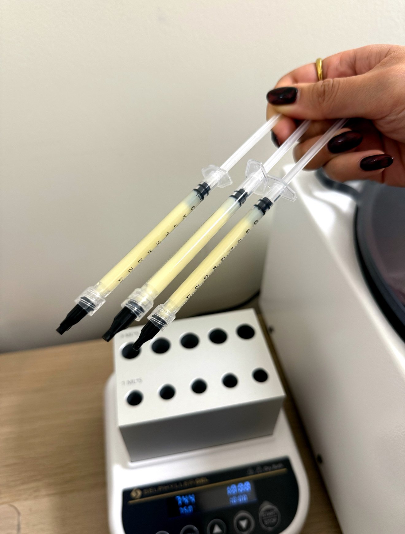 Hand holding three syringes filled with yellowish liquid, near a laboratory device on a wooden surface.