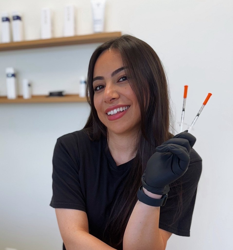 A young woman with long dark hair smiling and holding two used test tubes with orange caps in a laboratory setting.