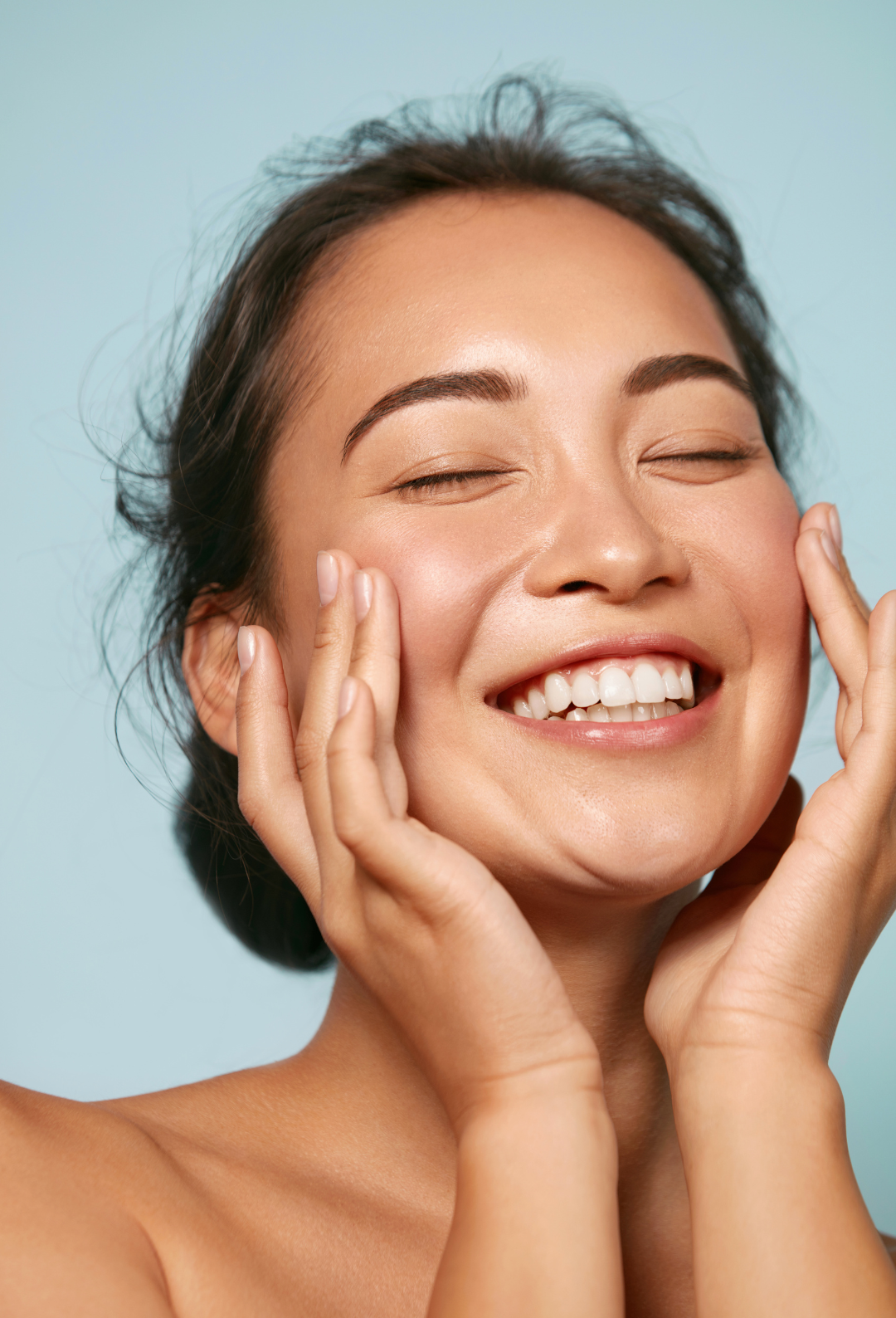 Close-up of a smiling woman with clear skin, closed eyes, and hands gently touching her face against a light blue background.