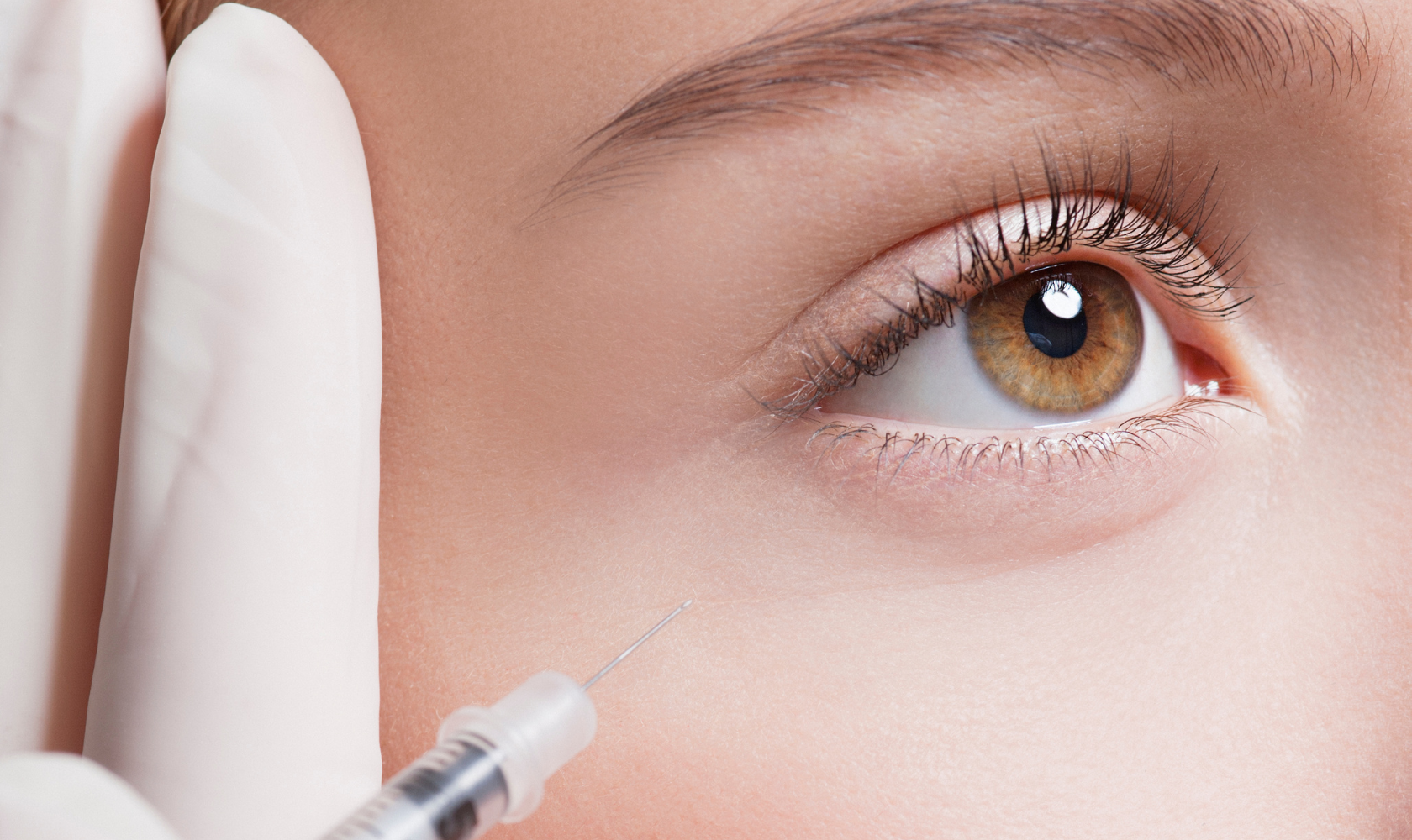 Close-up of a woman getting a cosmetic cosmetic injection near her eye, with a healthcare professional wearing a white glove holding a syringe.
