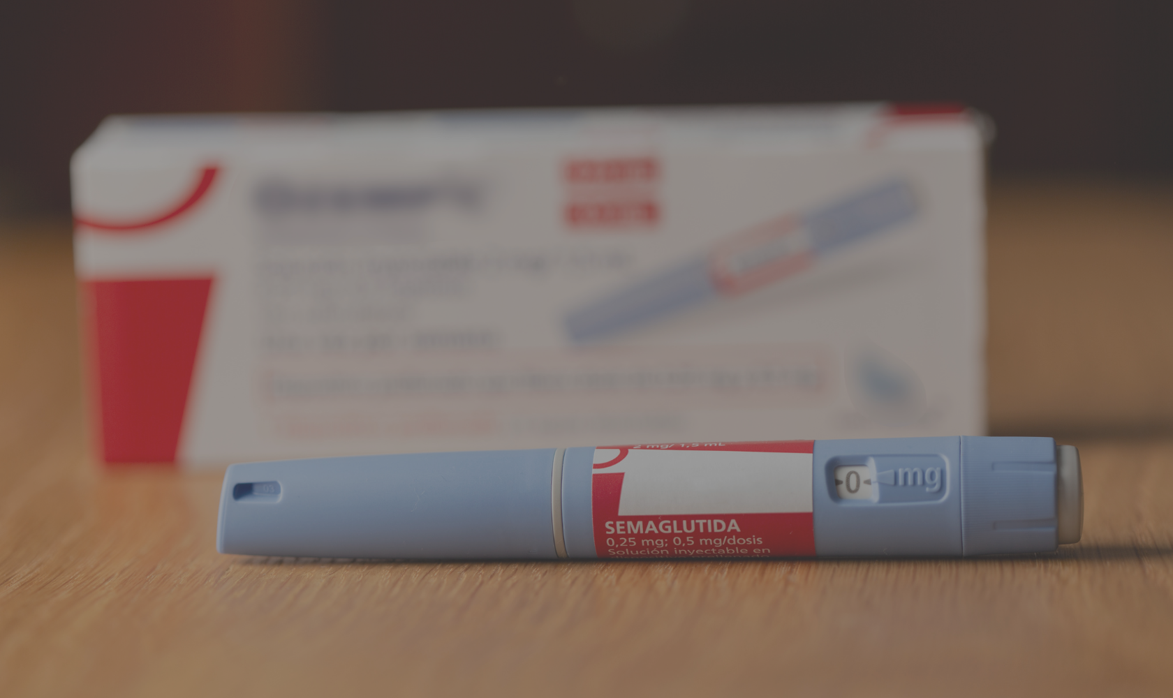 A medology pen with a red and gray label reading 'SEMAGLUTIDA 0.25 mg: 0.5 mg/dosis' rests on a wooden surface in front of its white box packaging with red and gray accents.
