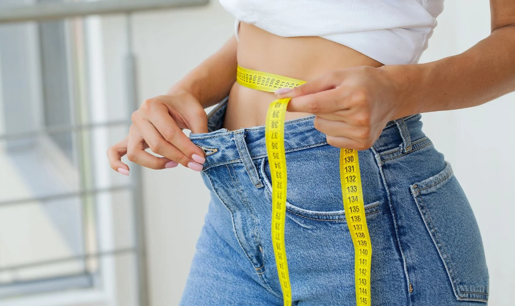 Person measuring waist circumference with a yellow tape measure, wearing blue jeans and a white top.