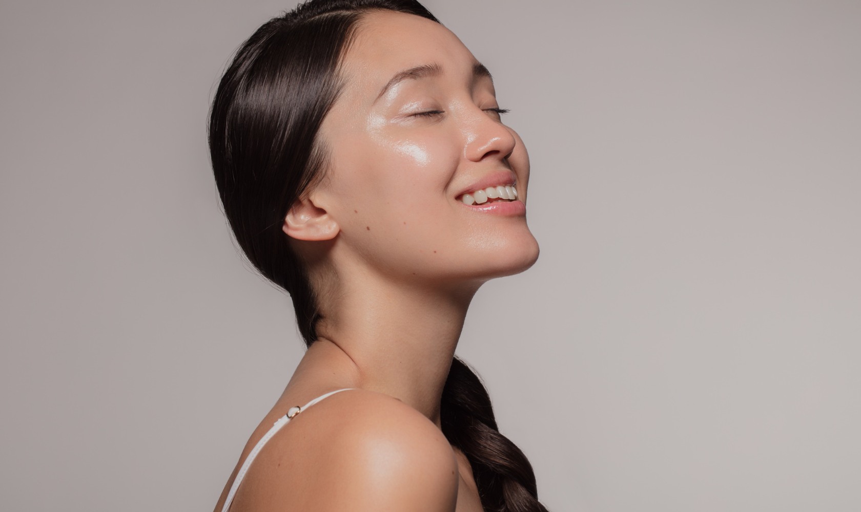 A woman with long dark hair smiling with her eyes closed, showing clear, radiant skin against a plain gray background.