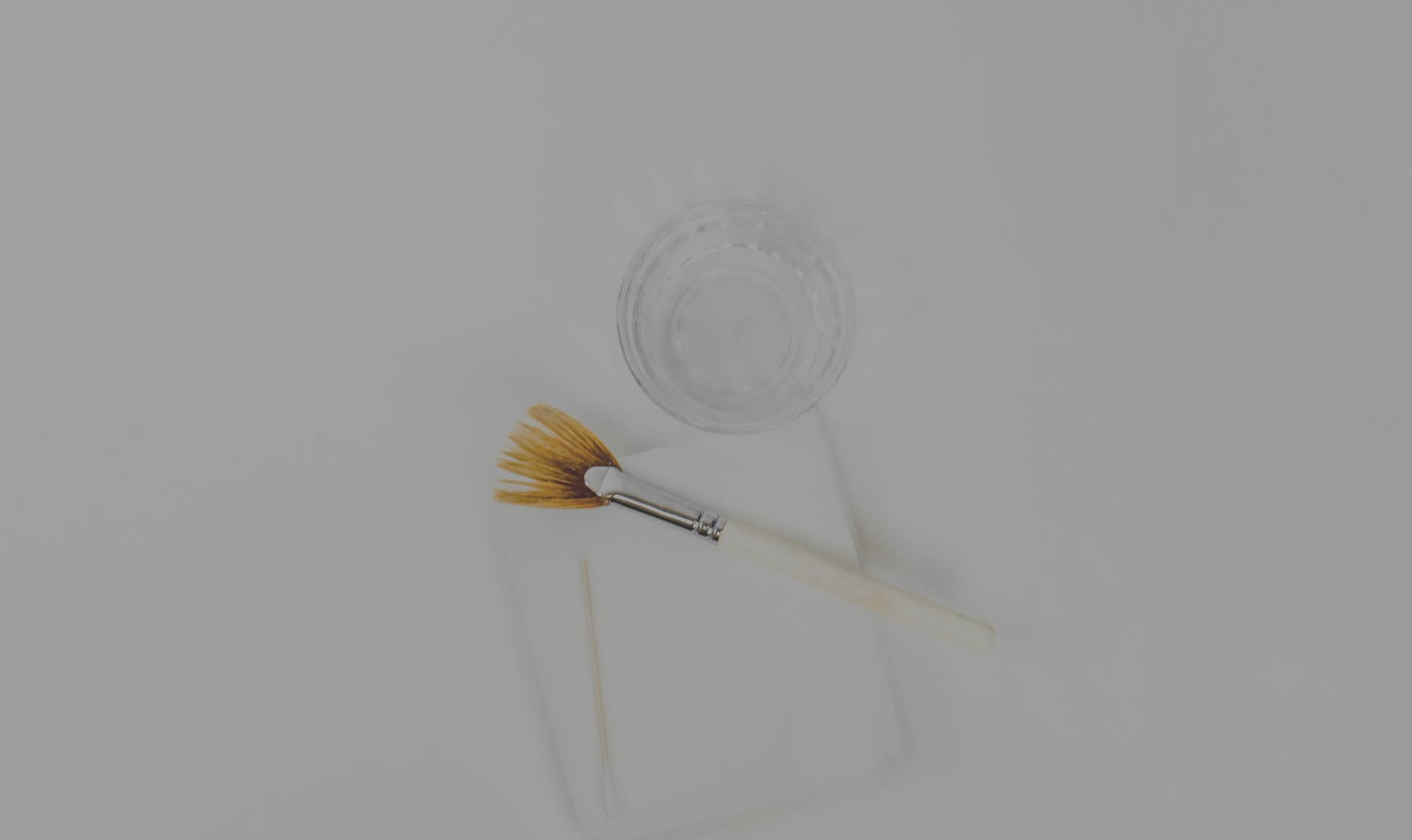 A glass of water, a makeup brush, and a spatula on a white surface.