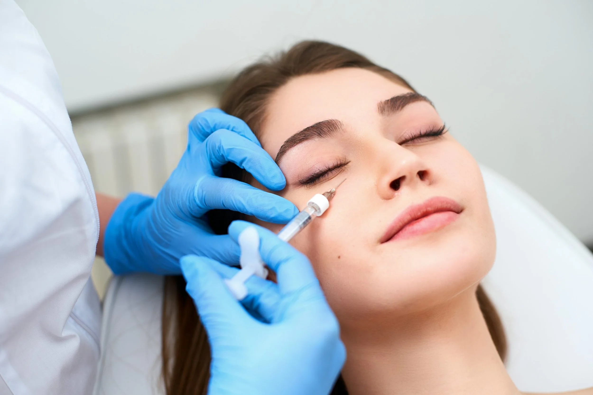 A woman receiving a cosmetic injection near her eye from a healthcare professional wearing blue gloves.