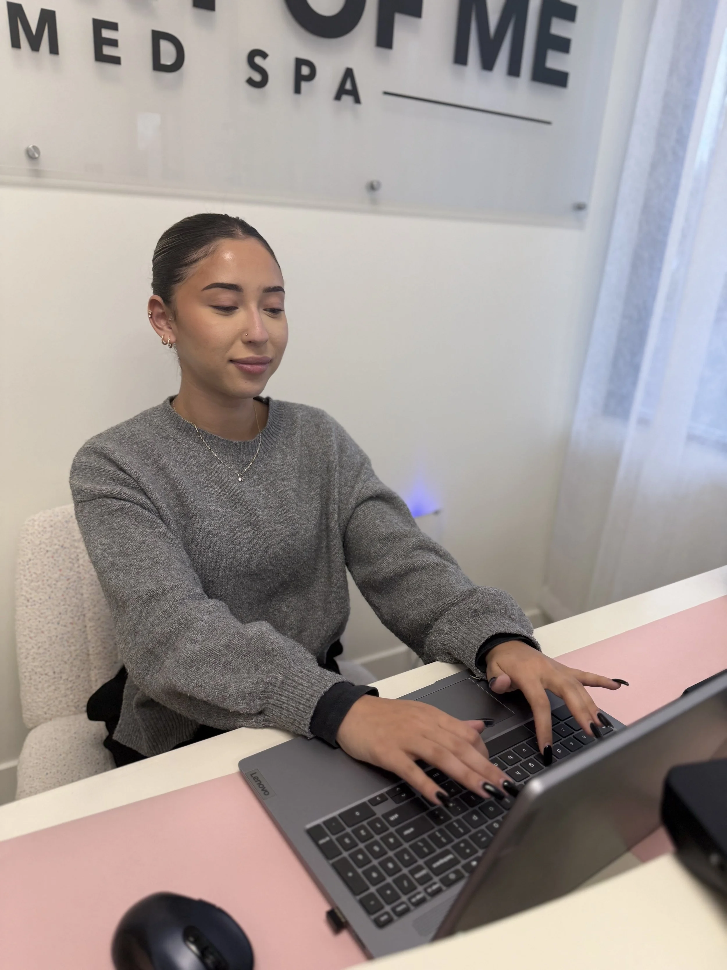A young woman with dark hair pulled back, wearing a gray sweater, working on a laptop at a desk in a wellness or spa center. Behind her, a wall displays the sign 'MED SPA'.