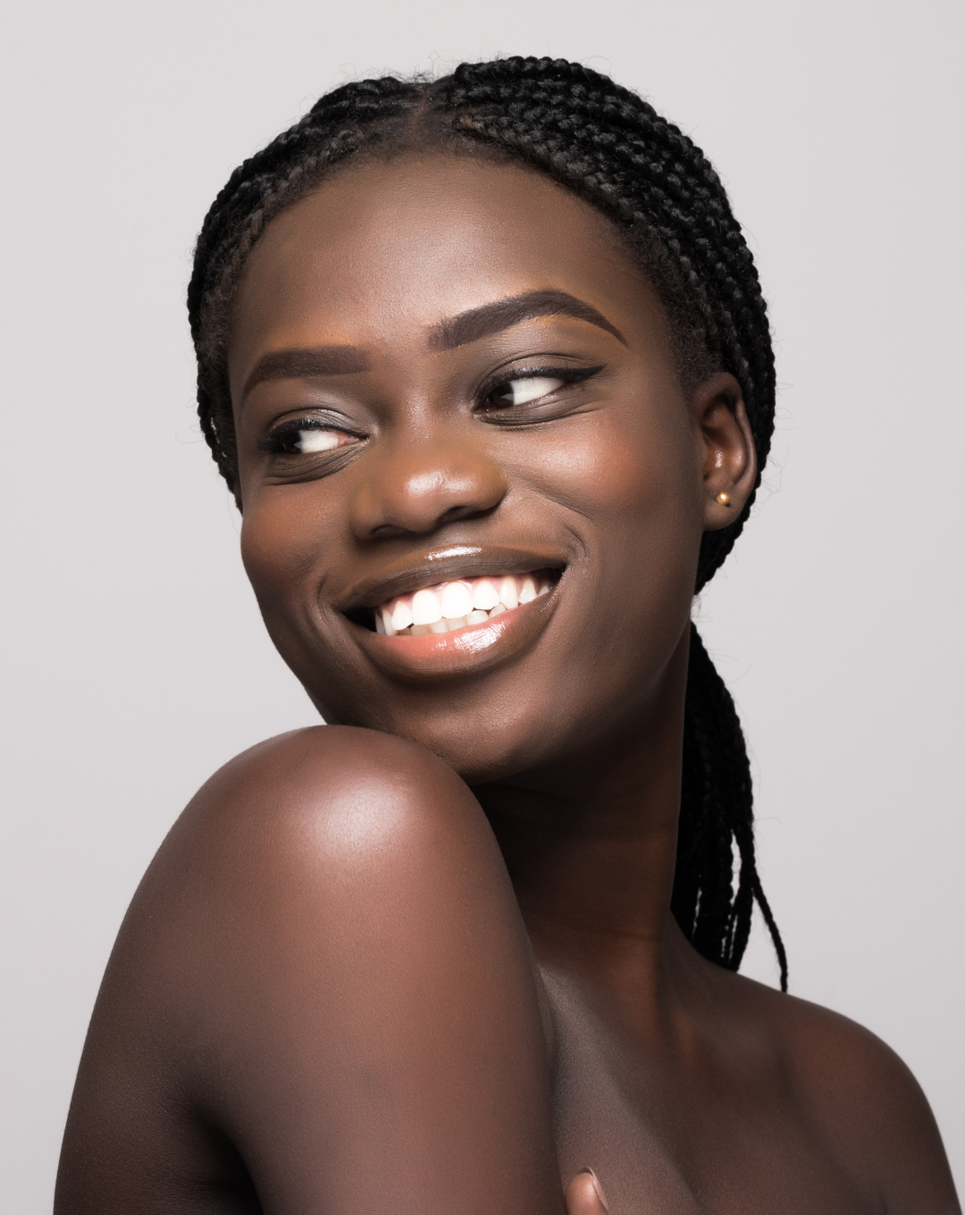 Close-up portrait of a smiling Black woman with braided hair, showing her shoulder and face, against a plain background.