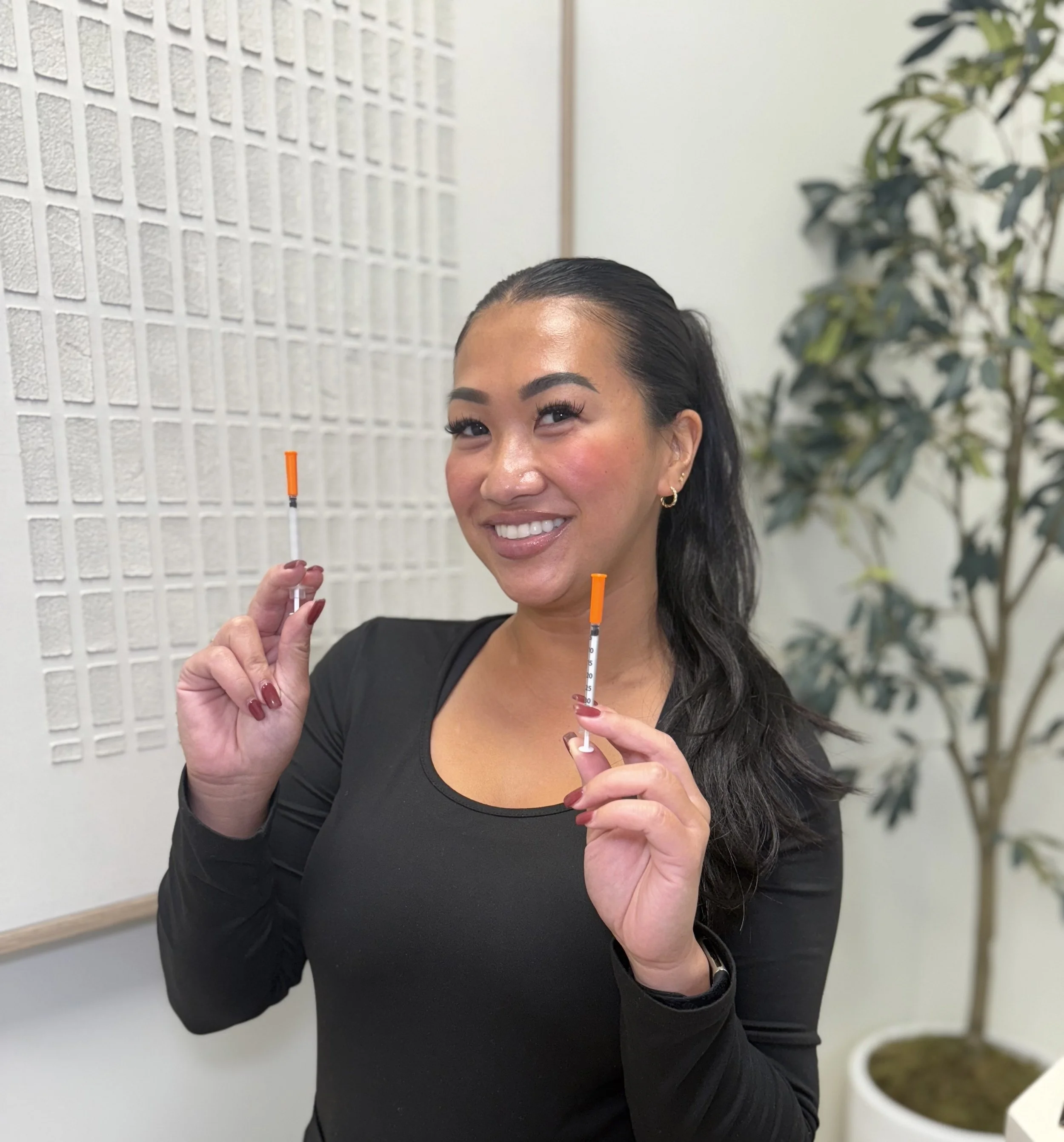 A woman with dark hair and earrings smiling, holding two syringes with orange caps, standing indoors next to a white wall and a plant.
