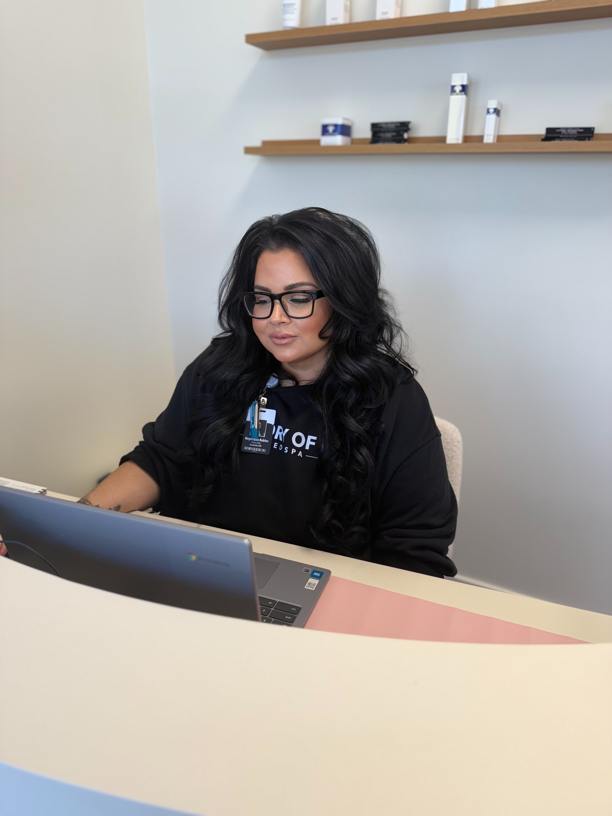 Woman with long black hair and glasses working on a laptop in an office, with shelves and products on the wall behind her.