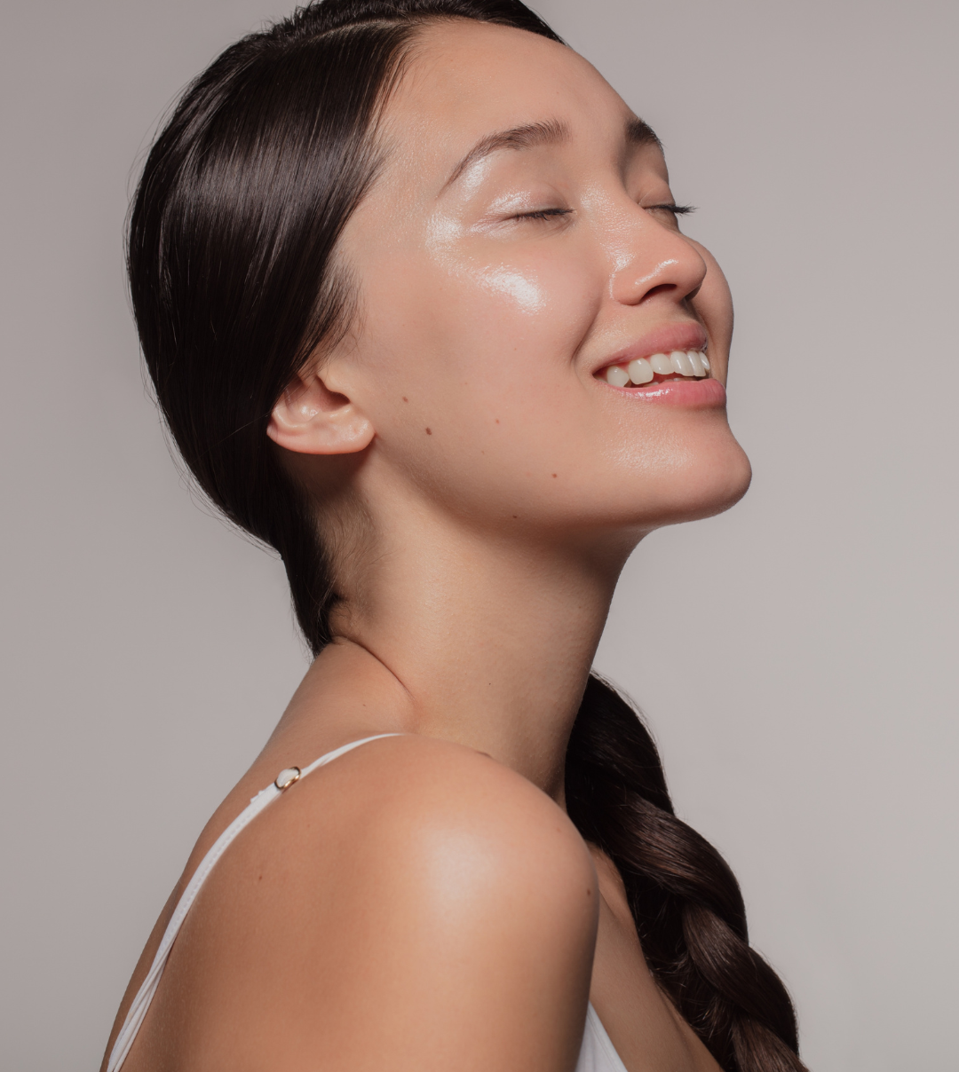 Close-up of a young woman with long brown hair styled in a braid, smiling with her eyes closed, wearing a white tank top, and having clear, glowing skin.