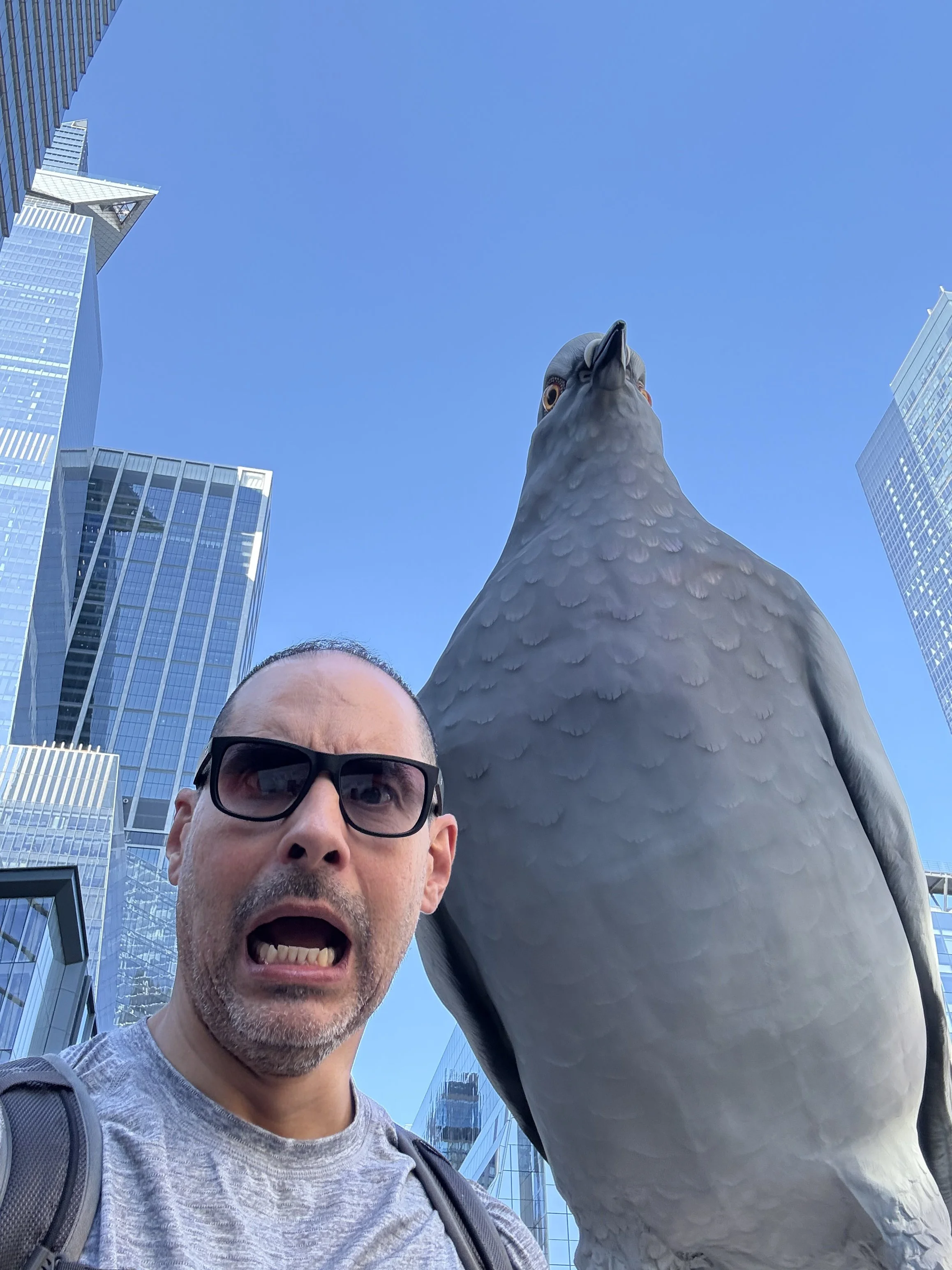 A man with glasses and a gray shirt taking a selfie next to a large pigeon statue in an urban setting with tall glass skyscrapers in the background on a clear day.