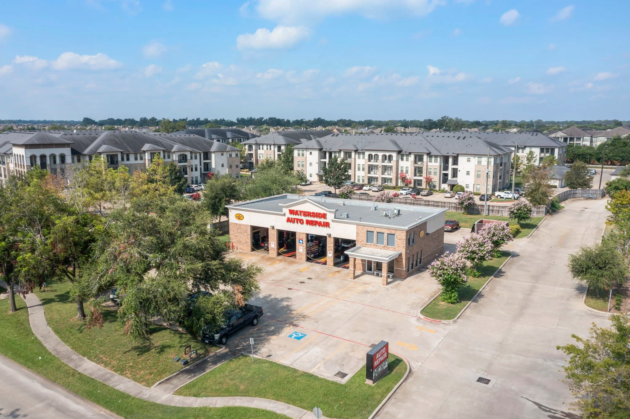 A parking lot in front of a multi-story apartment complex with a water-side auto repair shop in the foreground. Trees and a clear blue sky with scattered clouds surround the area.