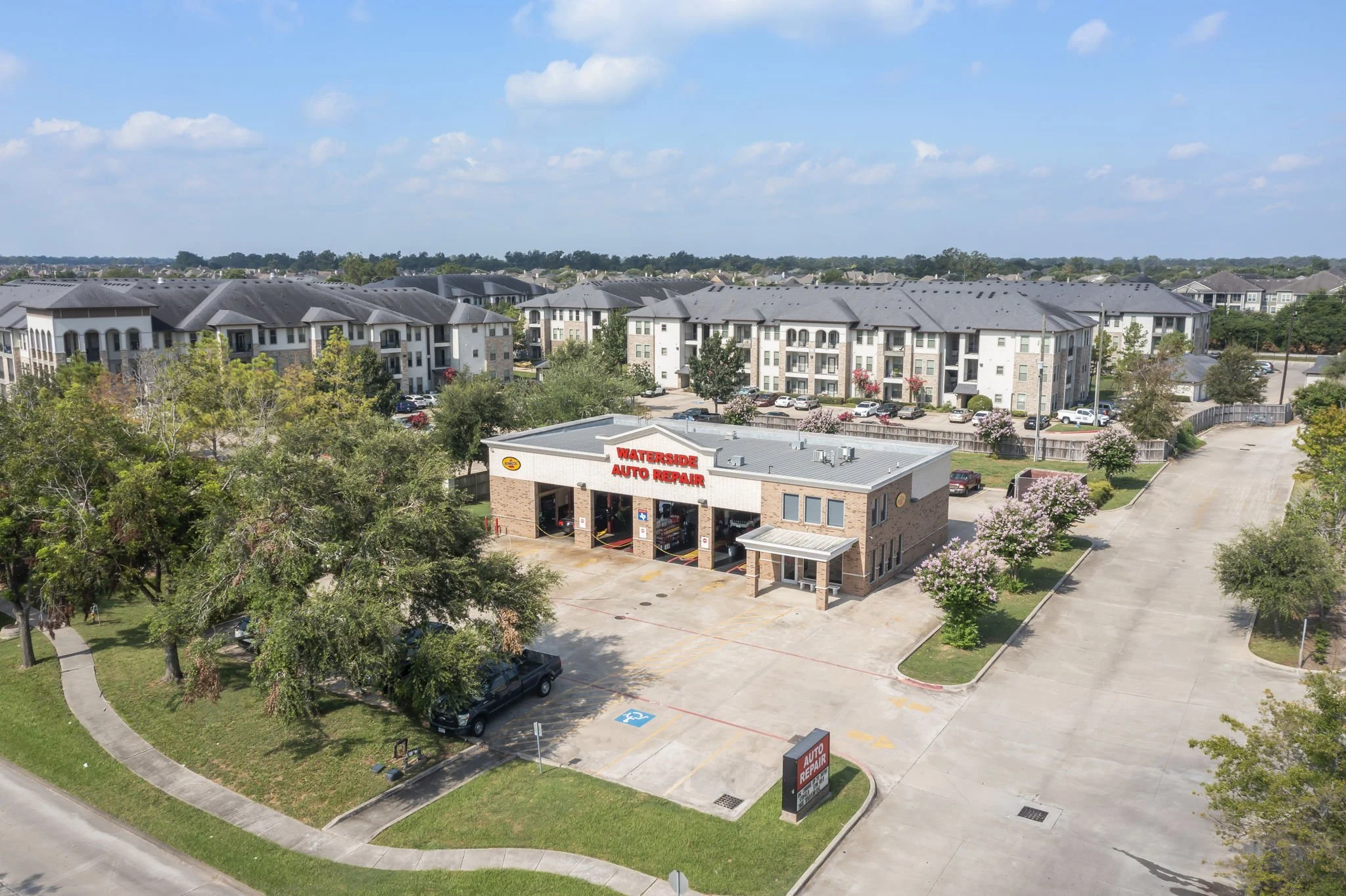 Aerial view of a residential apartment complex with multiple buildings, parking lots, and a nearby auto repair shop with a sign reading "WATERSIDE AUTO REPAIR." Trees and green spaces are interspersed throughout the area under a partly cloudy sky.