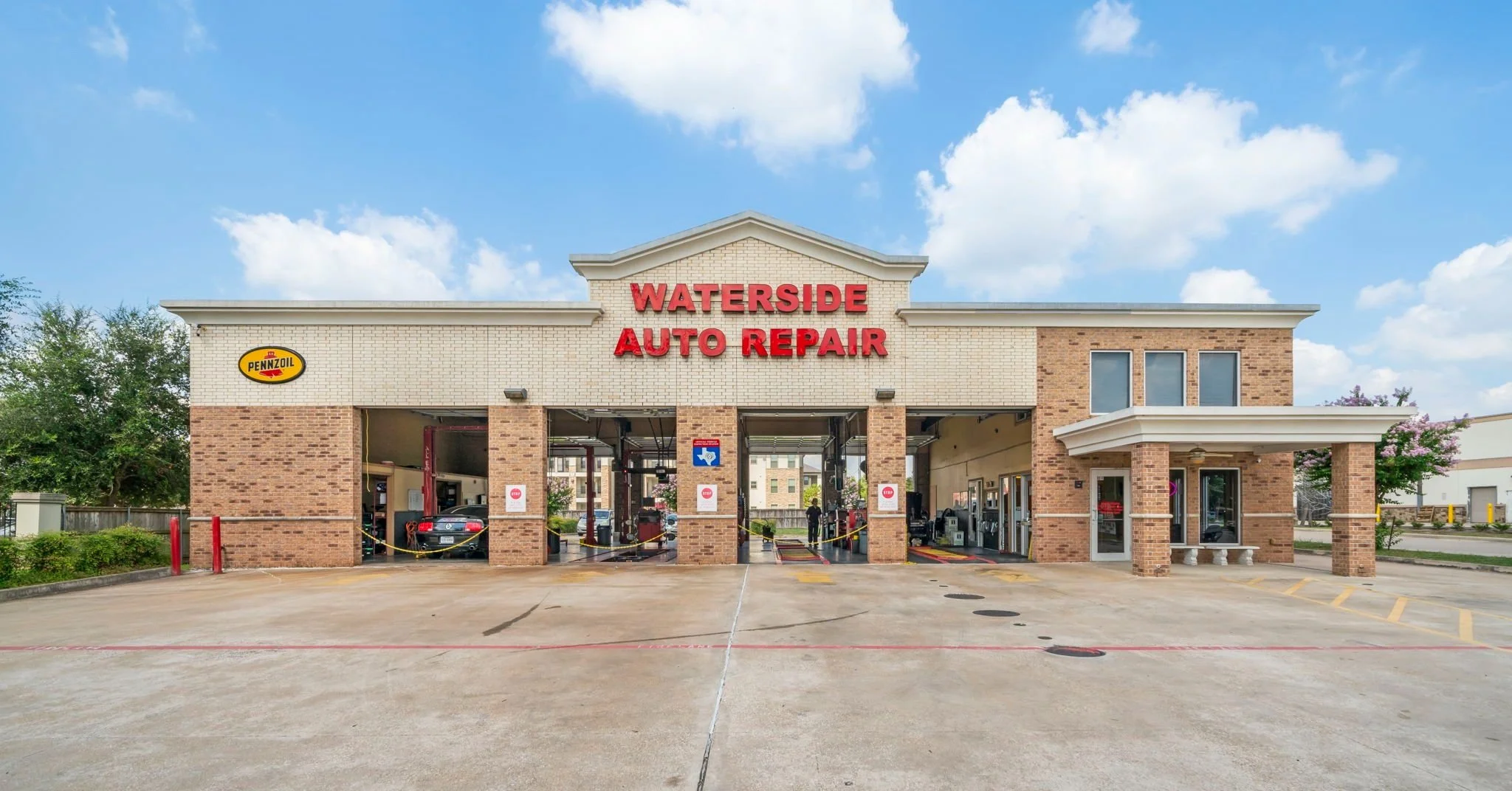 Exterior view of Waterside Auto Repair, a car repair shop with brick and white siding, multiple bays, and a large red sign. Pennzoil sign displayed on the building. Cars are inside the bays, and the parking lot is visible in the foreground with a few red posts and parking lines.