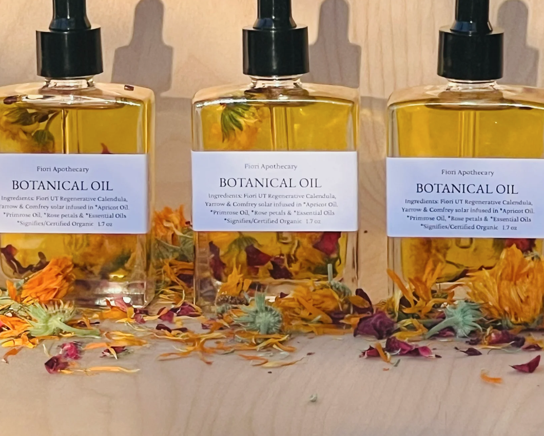 Three bottles of organic Botanical Oil surrounded by dried flowers, featuring calendula, yarrow, comfrey, apricot oil, primrose oil, rose petals, and essential oils at Kalm Sanctuary.