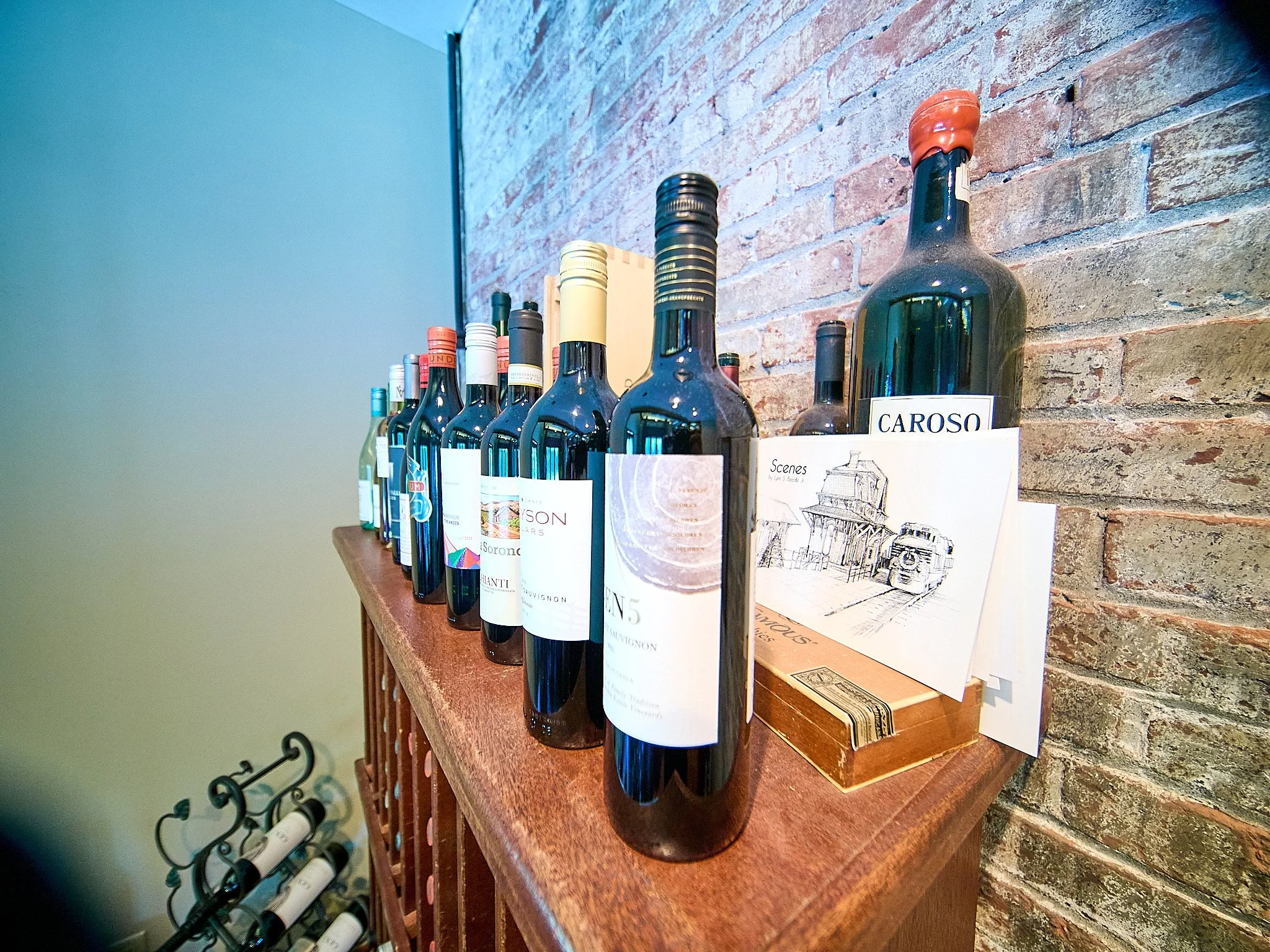 A row of wine bottles placed on a wooden cabinet against an exposed brick wall, with a small rack of wine bottles partially visible below.
