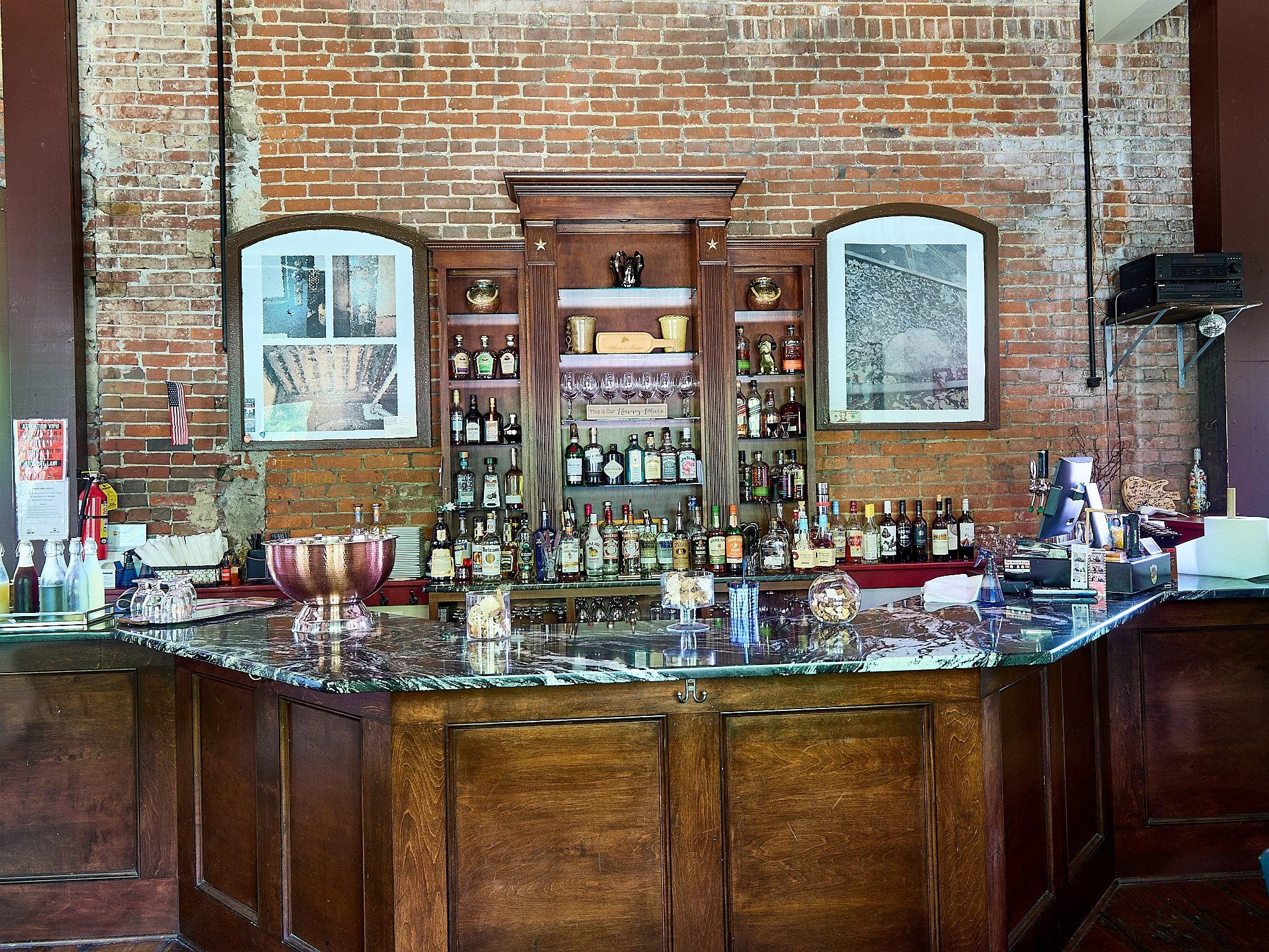 Bar with wooden cabinets, a marble countertop, and bottles of alcohol on the shelves, set against a brick wall with mirrors.
