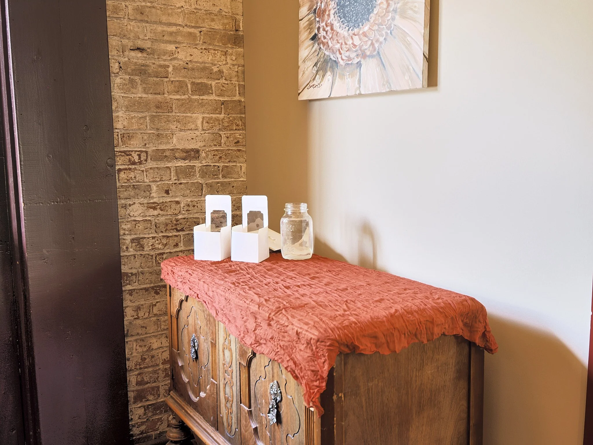A wooden sideboard draped with a crinkled red cloth, with two white tissue boxes and a glass jar on top, against a brick wall and a white wall with a painting.